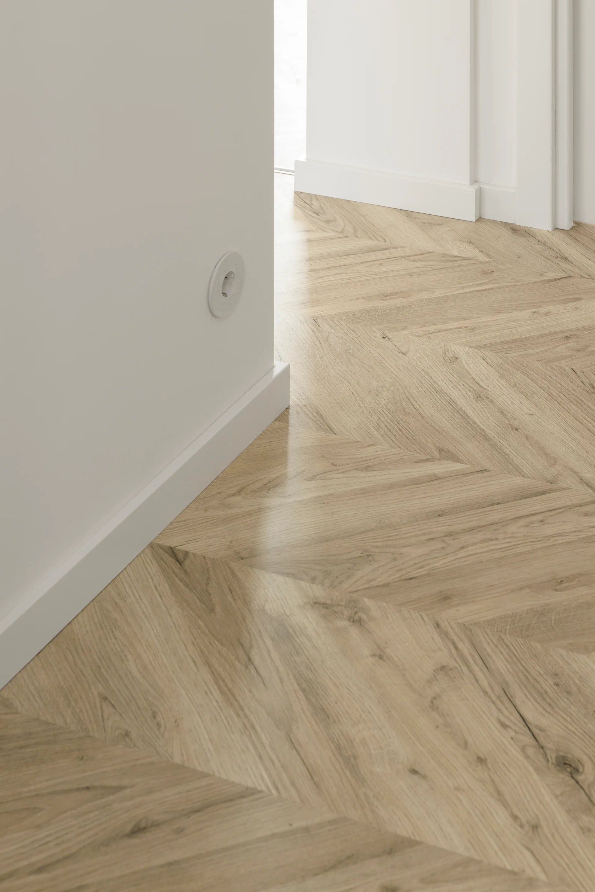 A close-up view of light wood herringbone flooring meeting a smooth white wall with crisp baseboard trim and a round electrical outlet.
