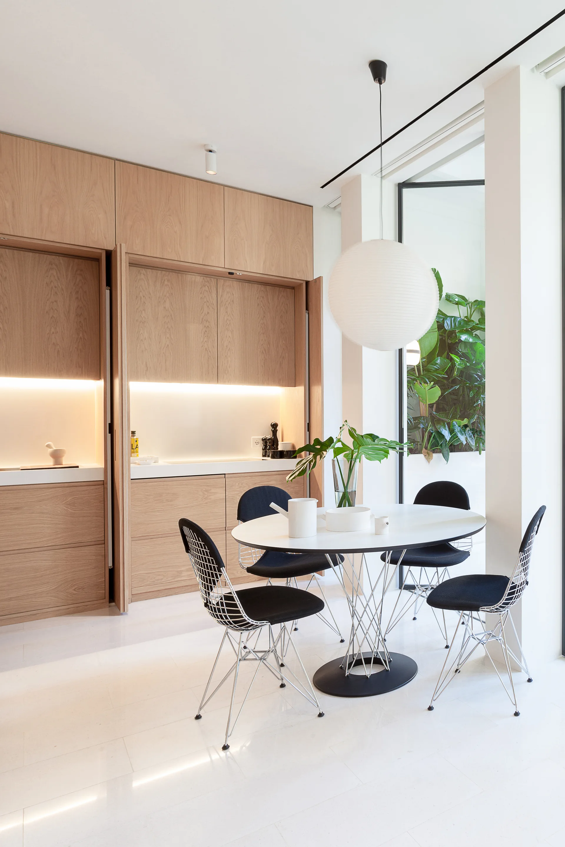 A minimalist dining area featuring an oval white table, black wire-frame chairs, a paper orb pendant light, and floor-to-ceiling light wood cabinets.