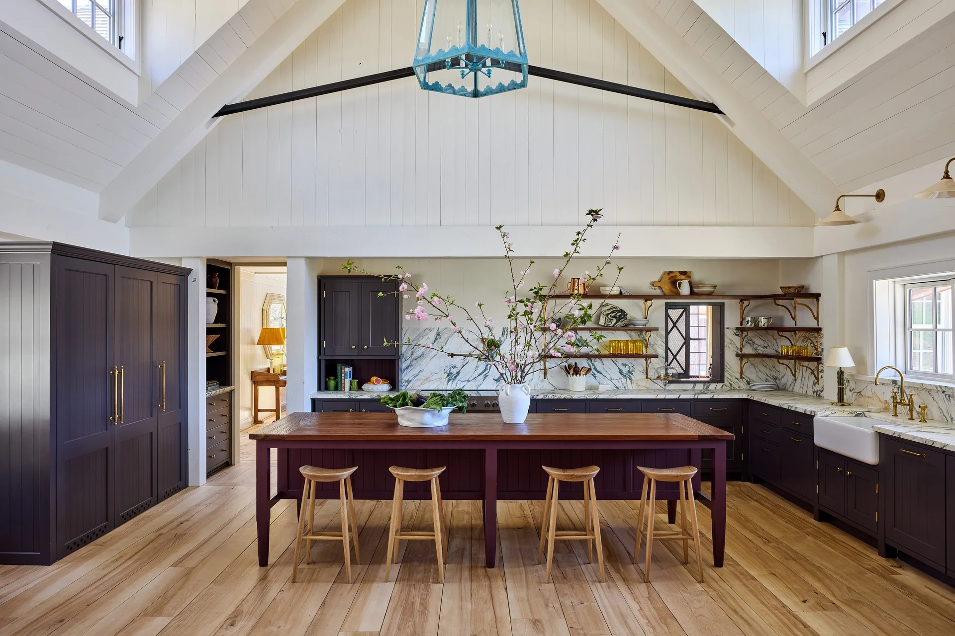 A high-ceilinged kitchen features dark purple cabinetry, a long wood-topped island with stools, marble countertops, and a white vaulted ceiling.