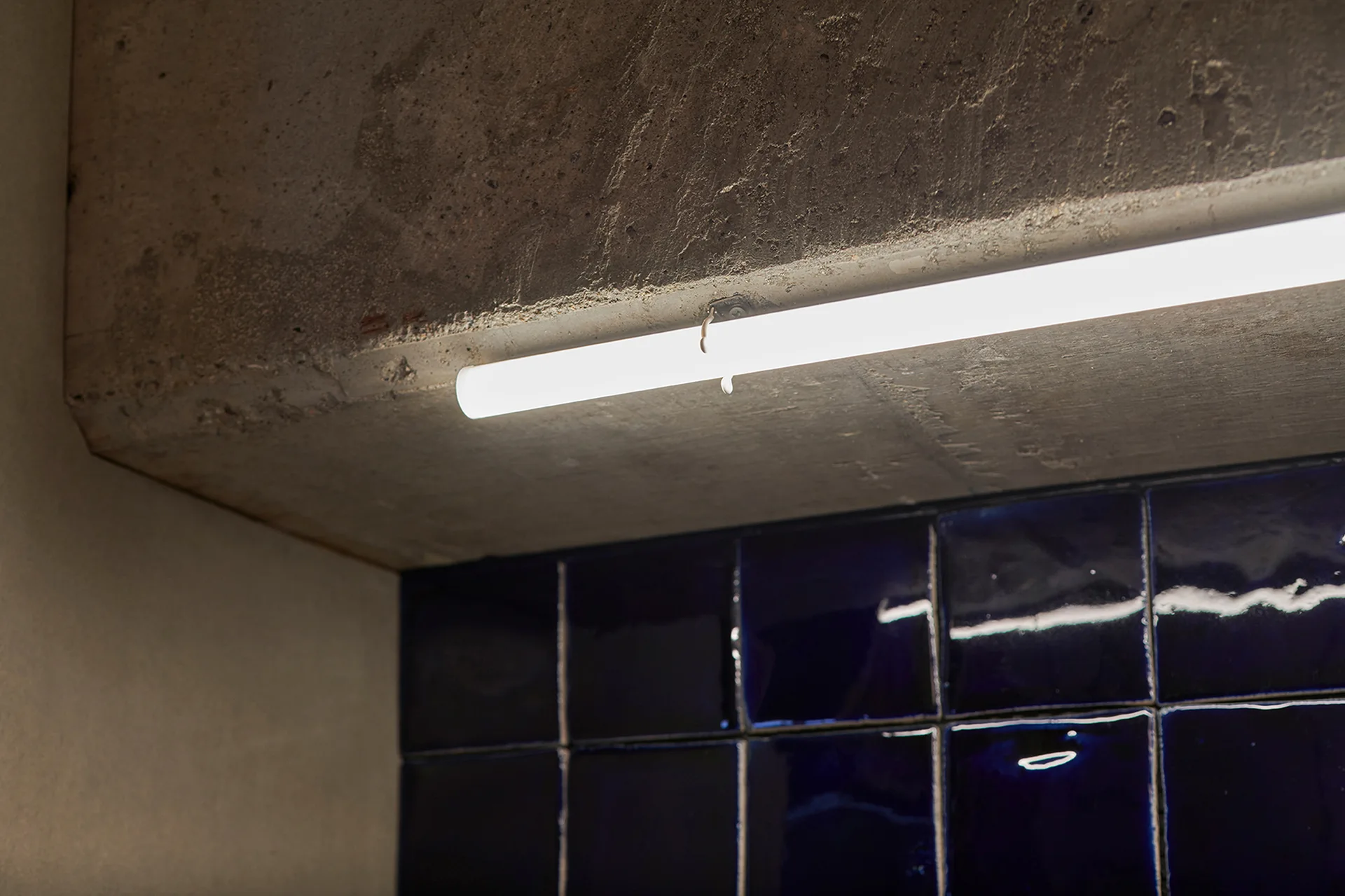 A glowing linear light fixture is mounted under a raw concrete beam above glossy dark blue square tiles.