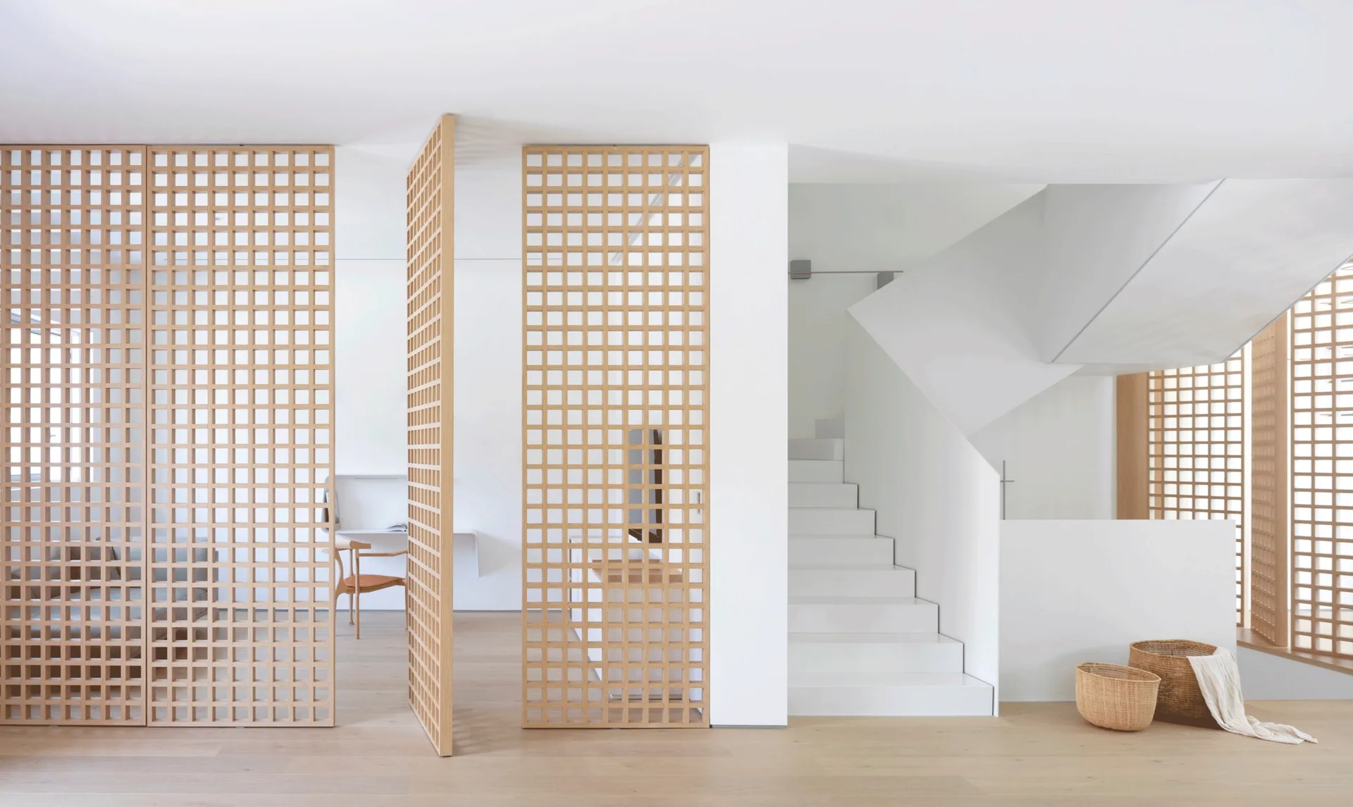 A minimalist interior featuring a white staircase, light hardwood floors, and large wooden grid partitions that act as room dividers.