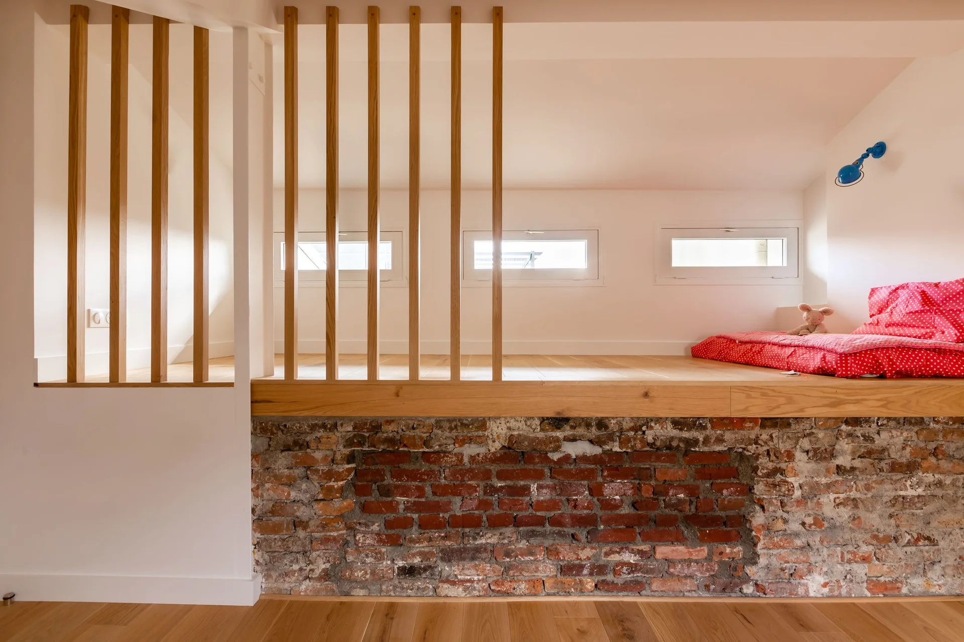A lofted bedroom platform features a vertical wood slat railing, light wood flooring, three small rectangular windows, and an exposed red brick base.