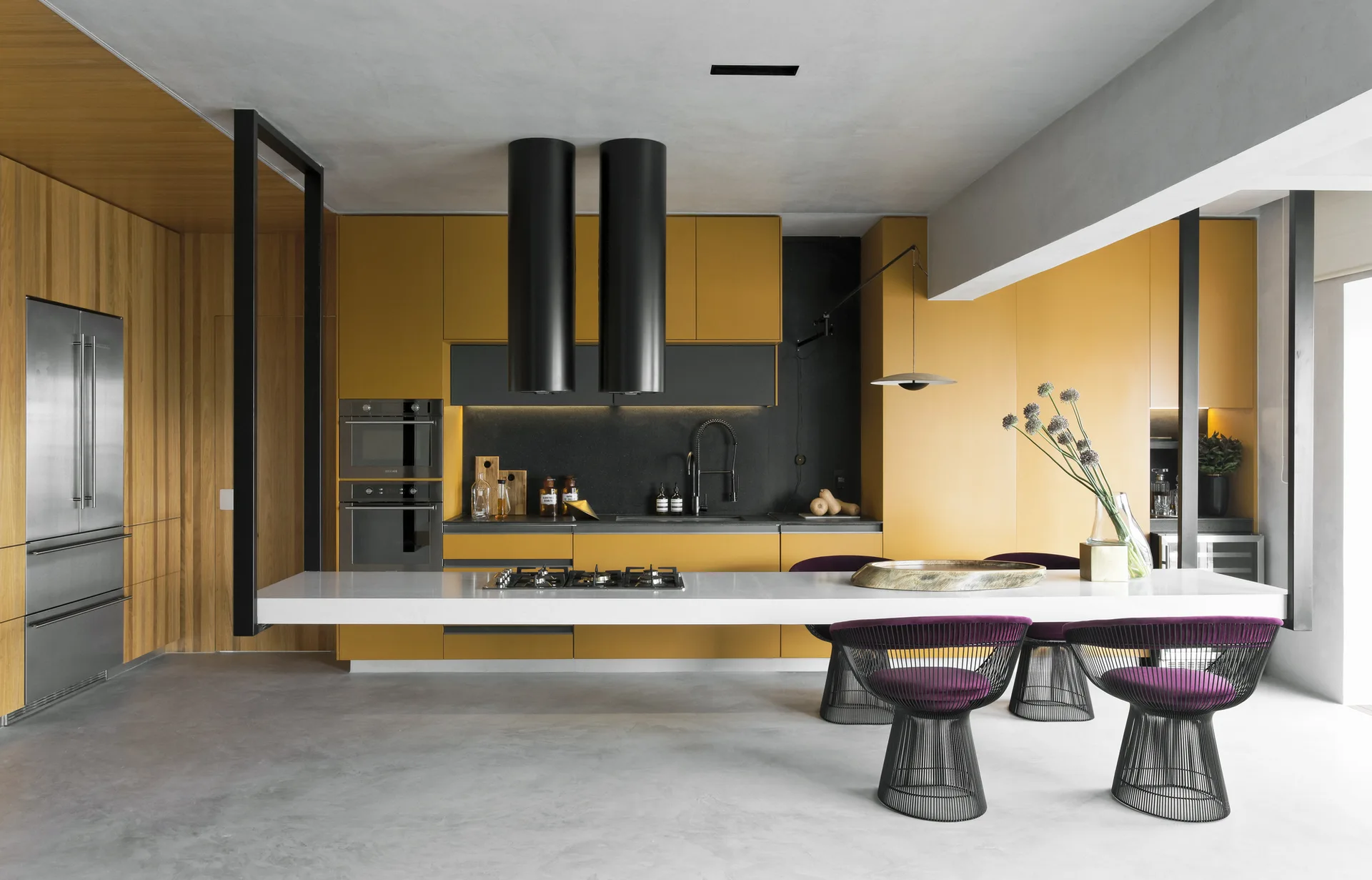 A modern kitchen with mustard yellow cabinetry, a long white cantilevered island, and black wire chairs with purple seat cushions.