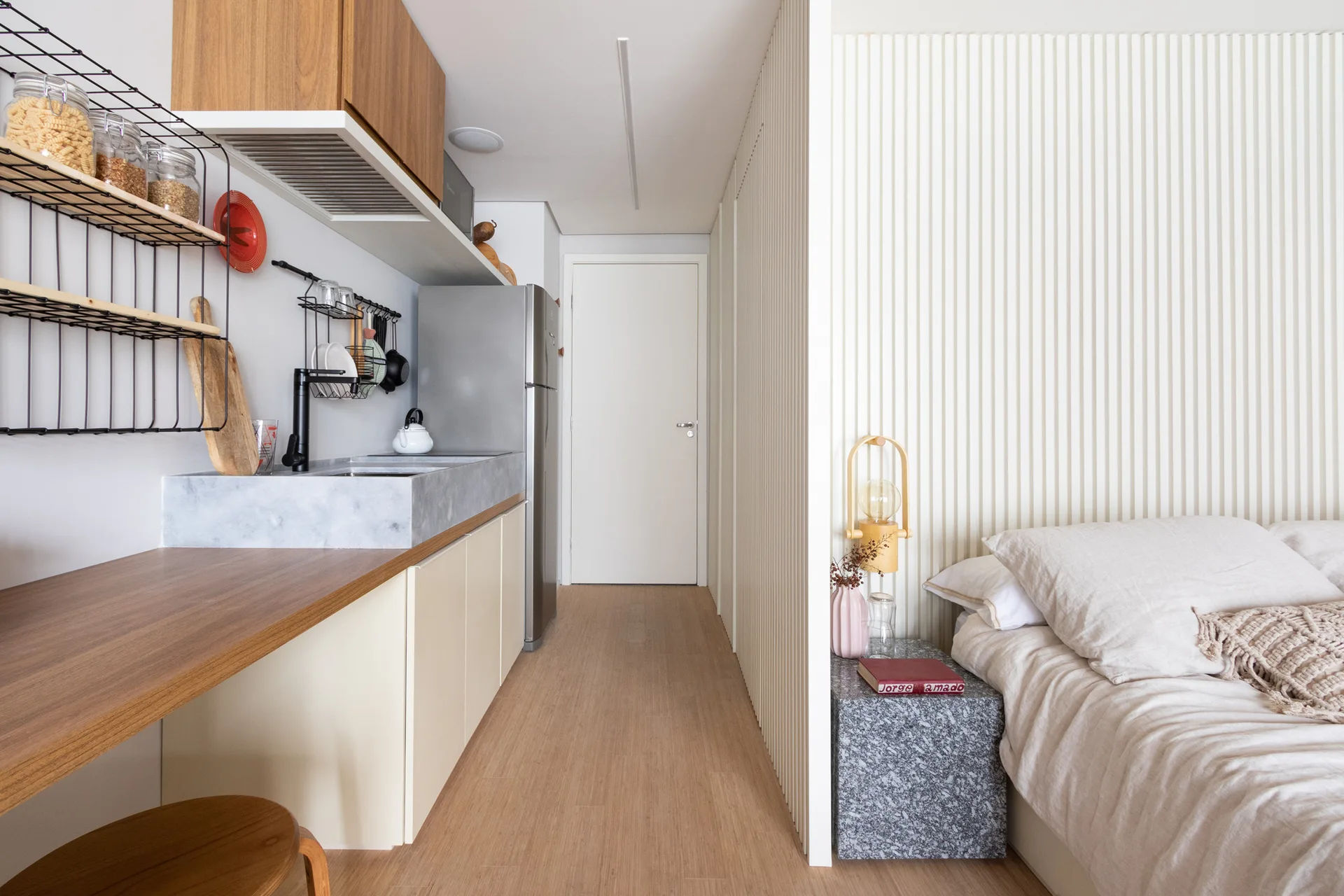 A compact studio apartment features a small kitchen with a stone sink next to a bed separated by a white slatted partition wall.