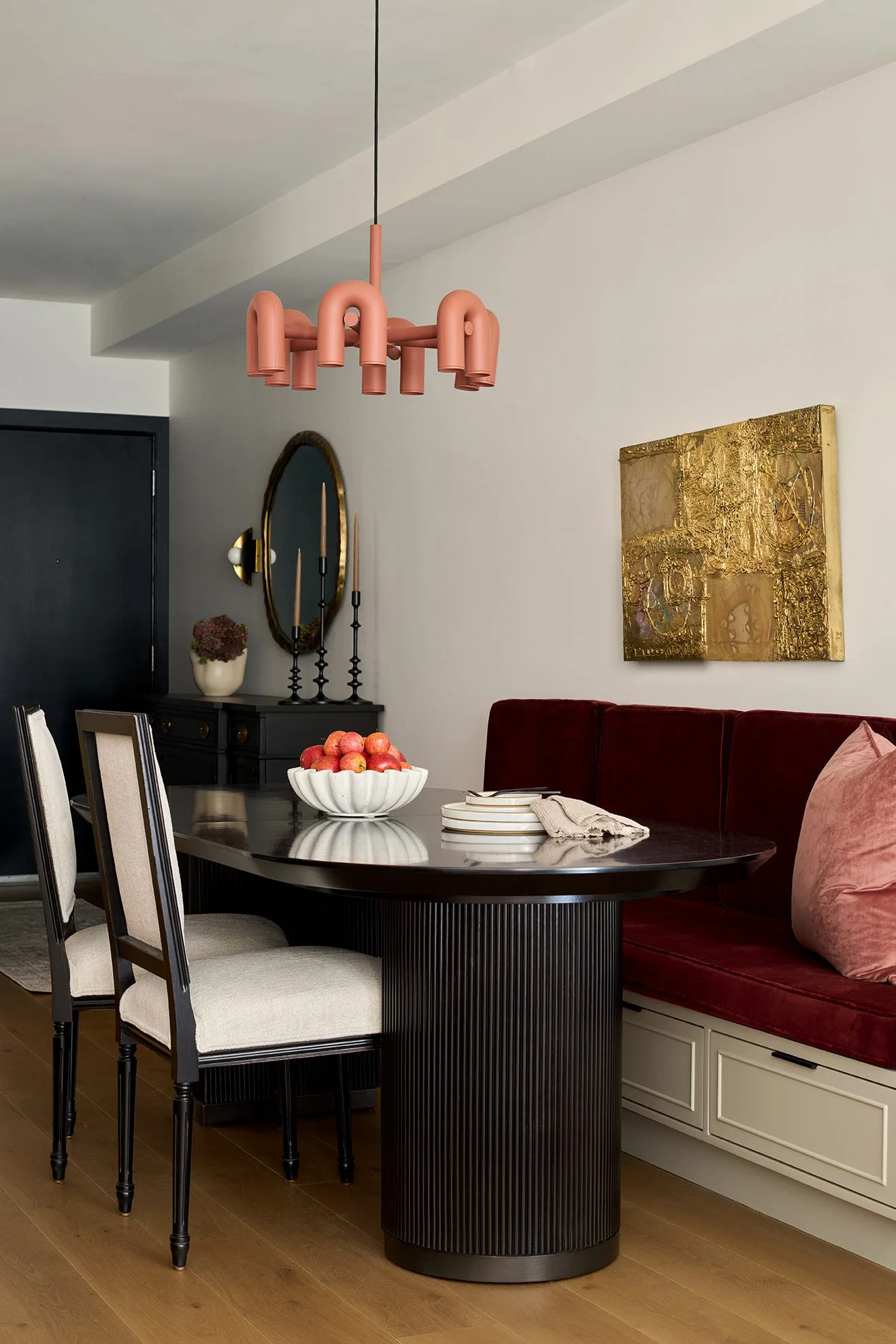 A modern dining area with a black oval pedestal table, a burgundy velvet banquette, and a sculptural pink arched pendant light.