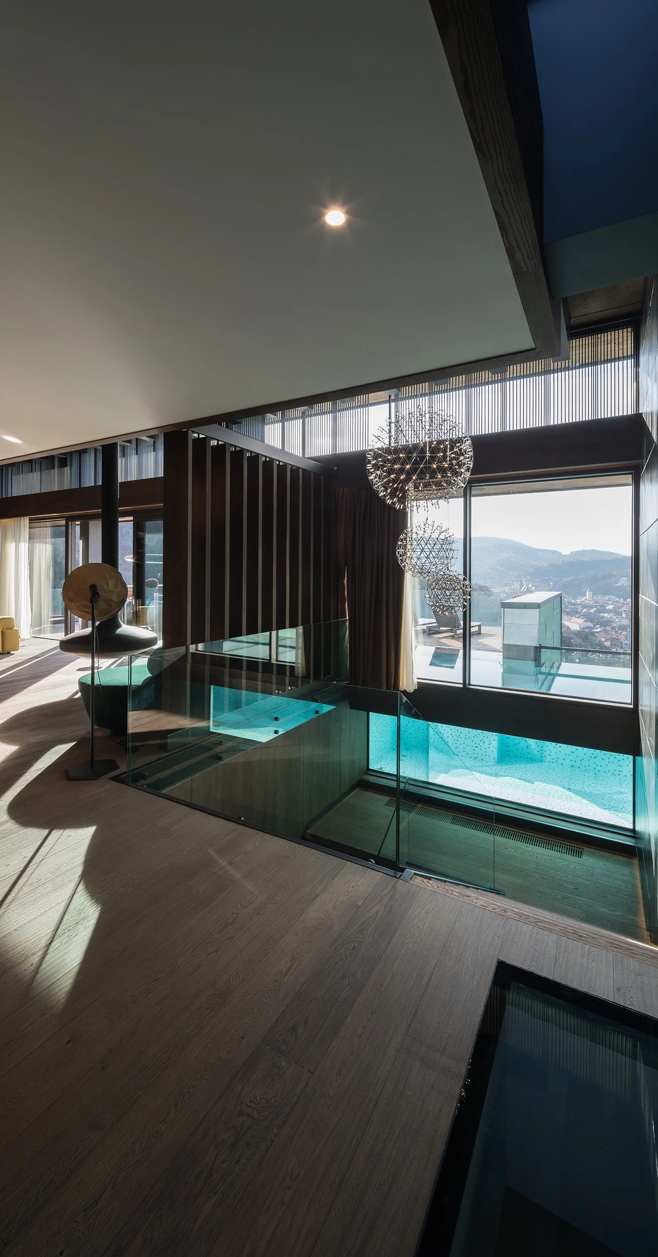 A modern split-level room with wood floors, glass railings overlooking an indoor pool below, and orb chandeliers hanging near a large window.
