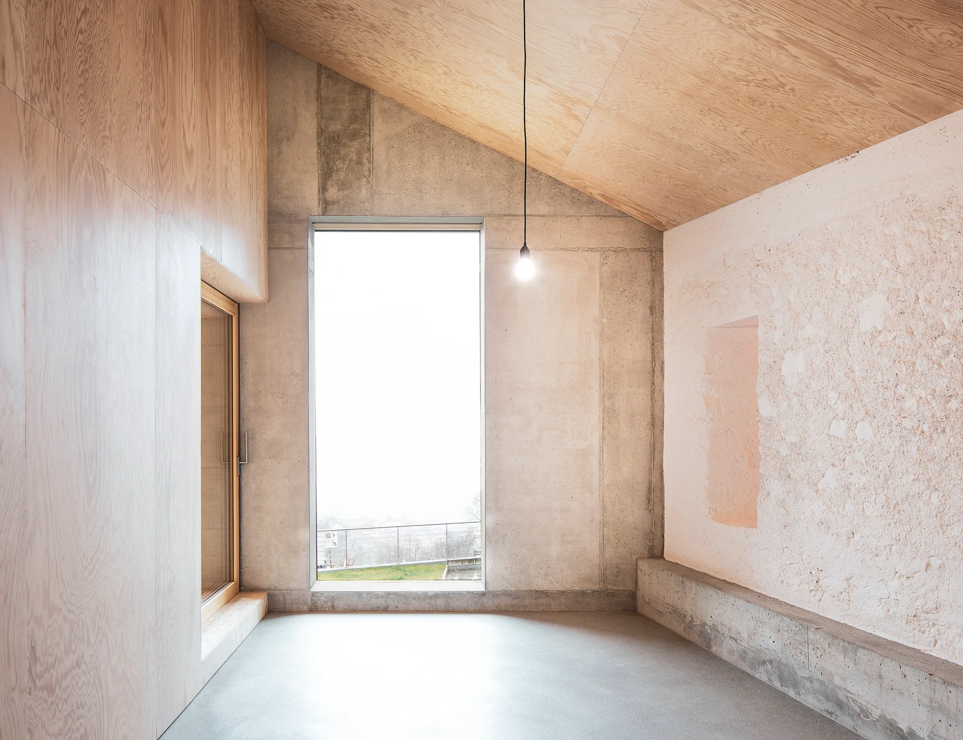 A minimalist interior room features a sloped wood-paneled ceiling, exposed concrete walls, a textured plaster wall, and a large central window.