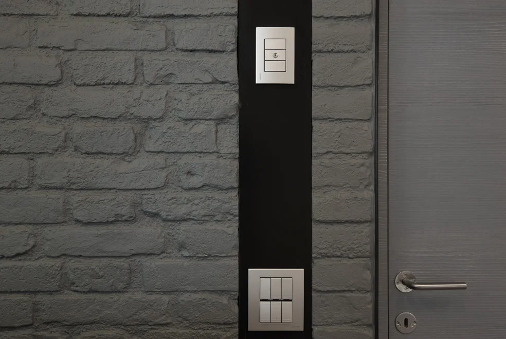 Close-up of a textured grey painted brick wall featuring a vertical black accent strip with two modern silver light switches next to a grey door.