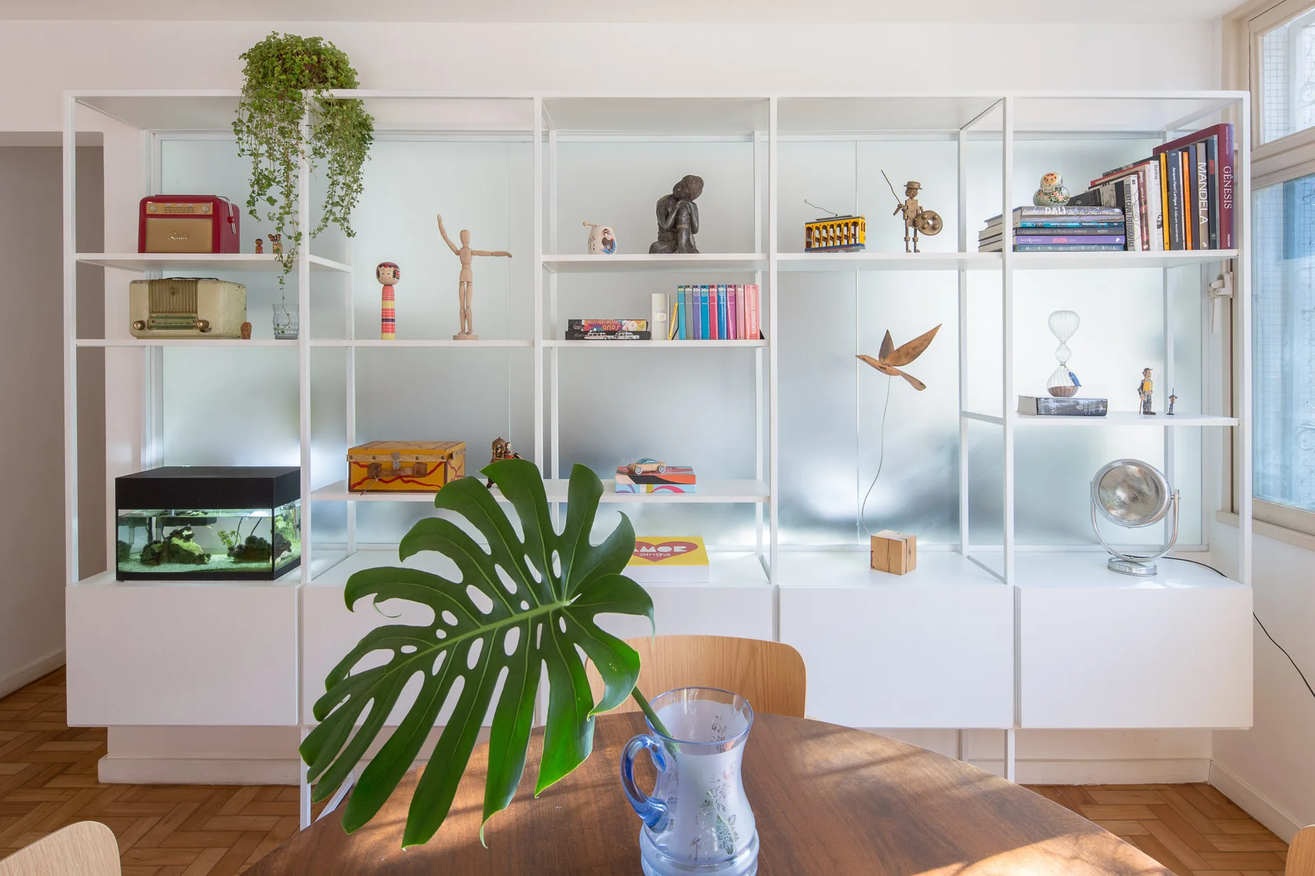 A white grid shelving unit displaying books and decor stands behind a round wooden dining table set with a blue glass pitcher holding a monstera leaf.