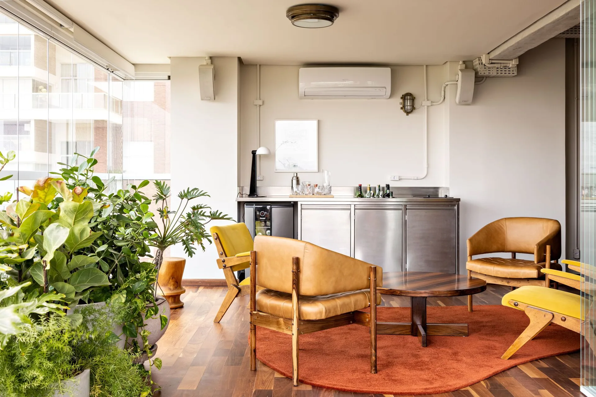 A sunlit indoor lounge features mid-century wood and leather chairs, a round wooden coffee table, a terracotta rug, and a stainless steel bar counter.