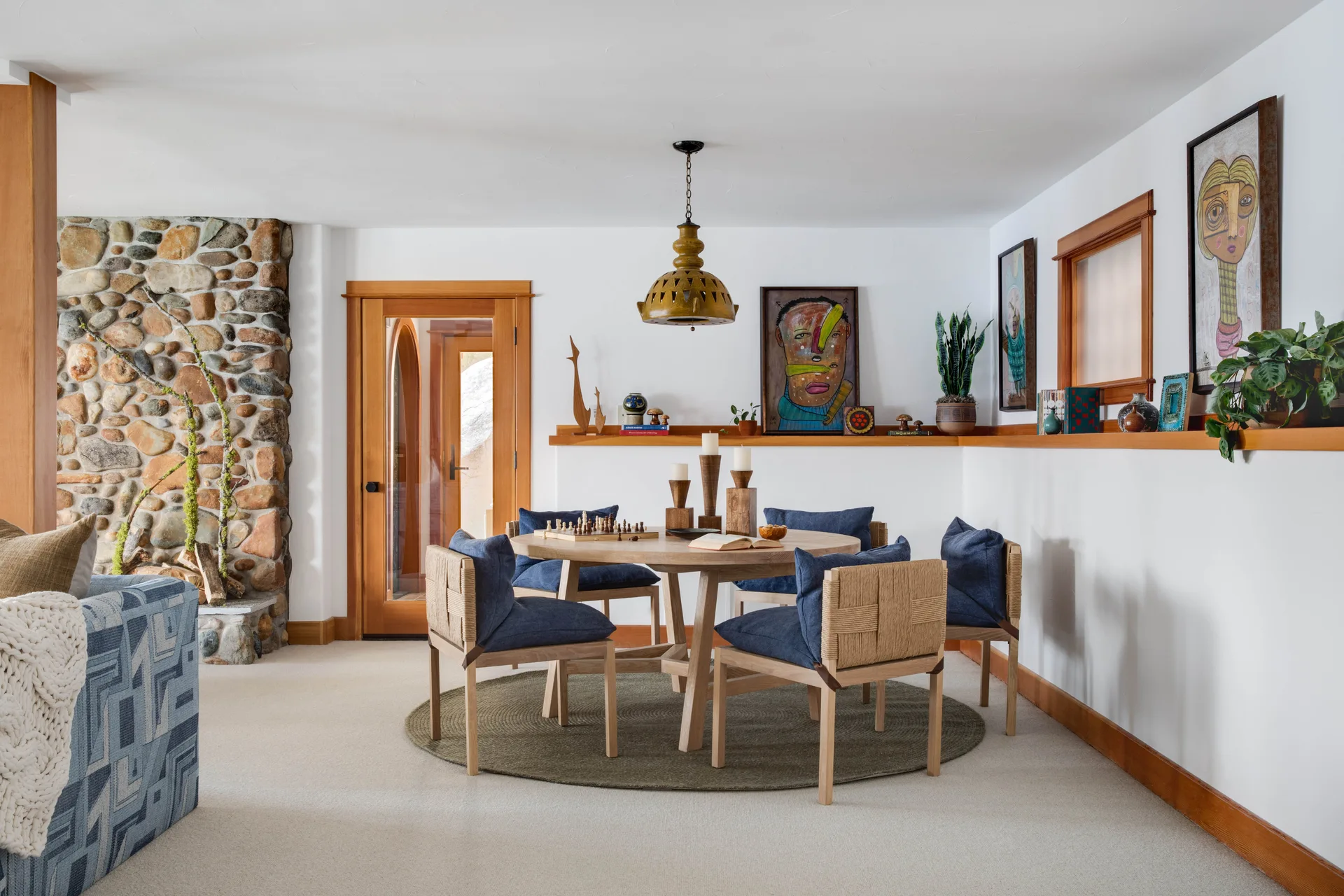 A round wooden dining table with blue chairs on a circular green rug, set against white walls with a continuous wood display shelf and a stone fireplace.