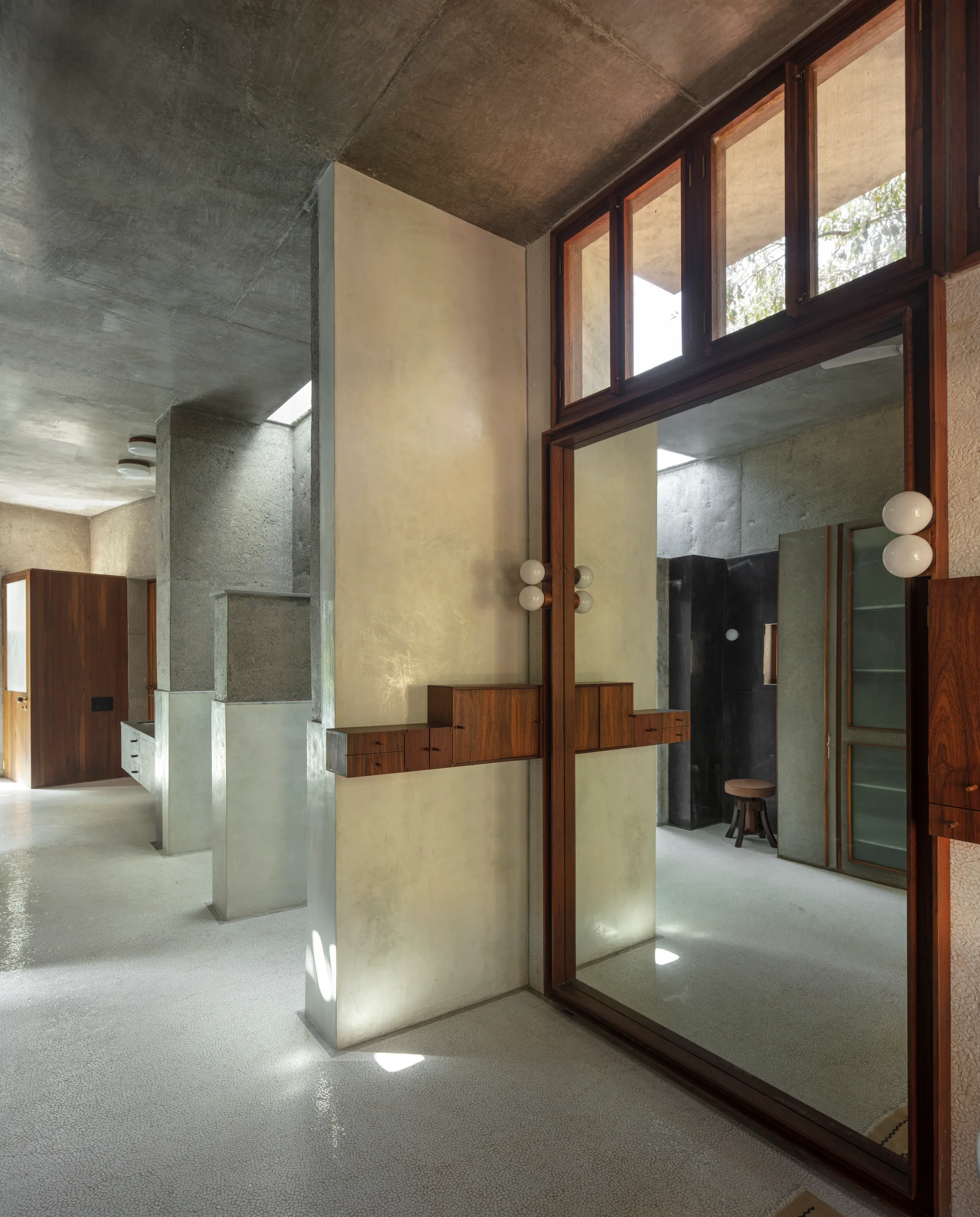 A minimalist concrete hallway features a large wood-framed mirror with integrated wall cabinets, light terrazzo flooring, and concrete walls.