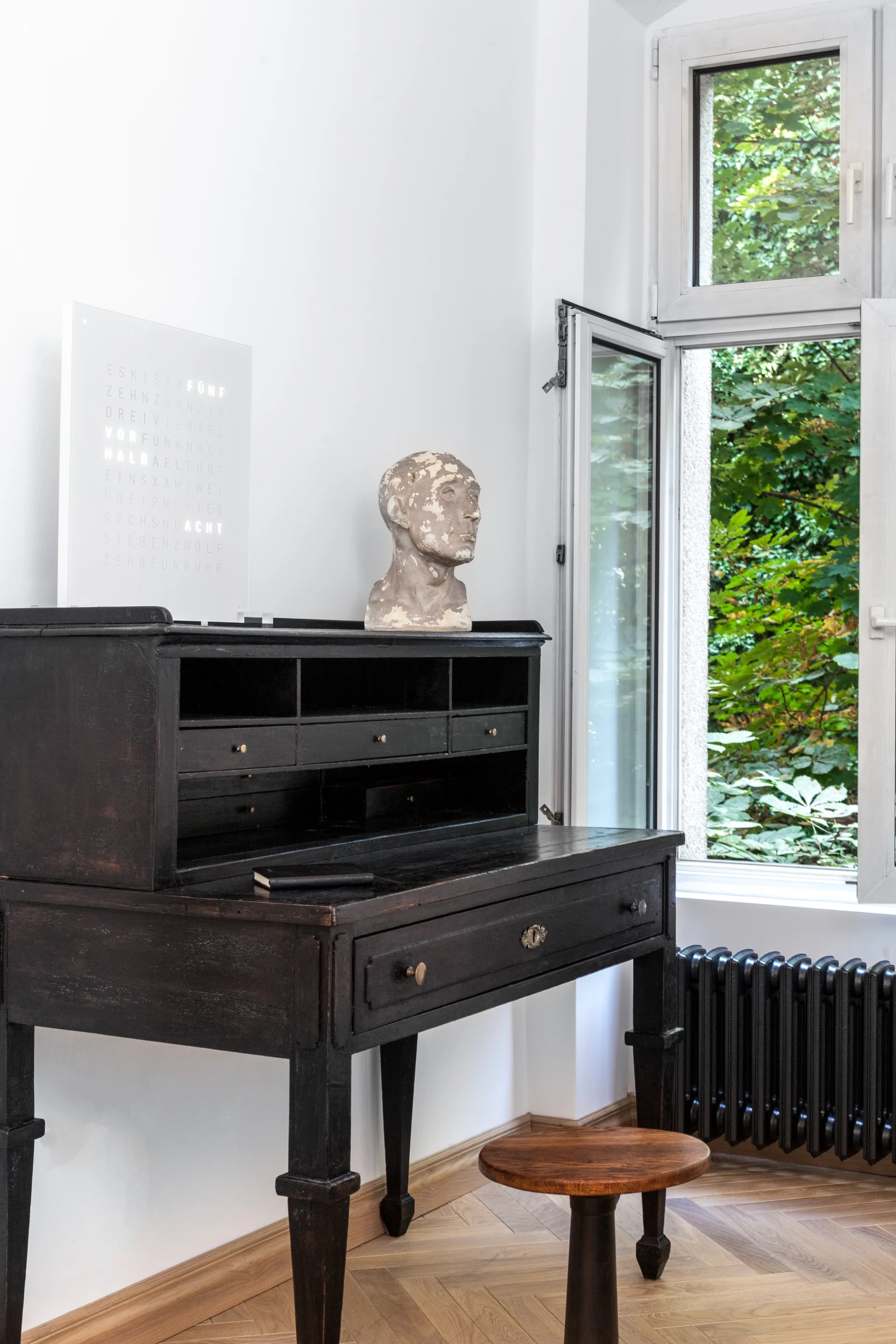 An antique black secretary desk decorated with a stone bust and word clock sits on herringbone wood floors next to an open window.