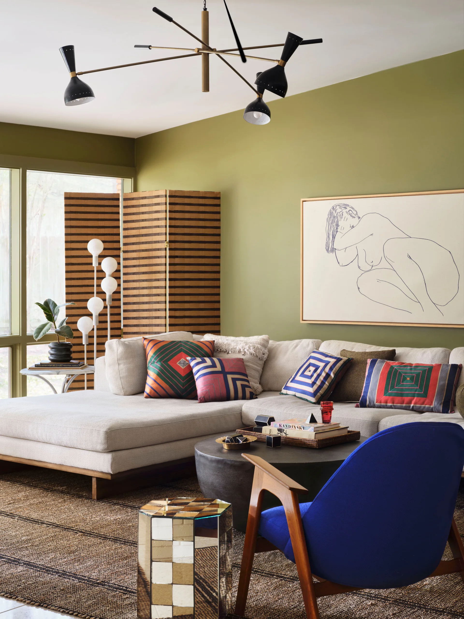 A living room with olive green walls features a cream sectional sofa, colorful geometric pillows, a blue accent chair, and a mid-century chandelier.