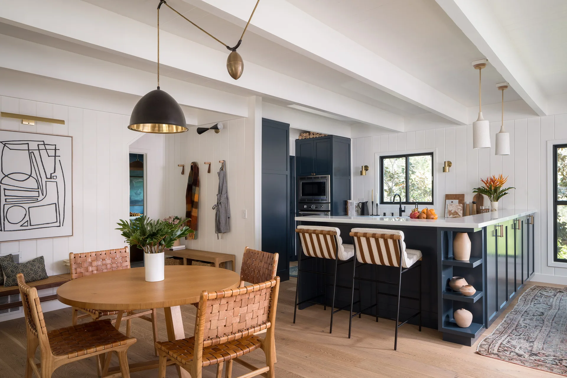 An open kitchen and dining area features a navy blue island with barstools, a round wooden dining table with woven leather chairs, and white paneled walls.