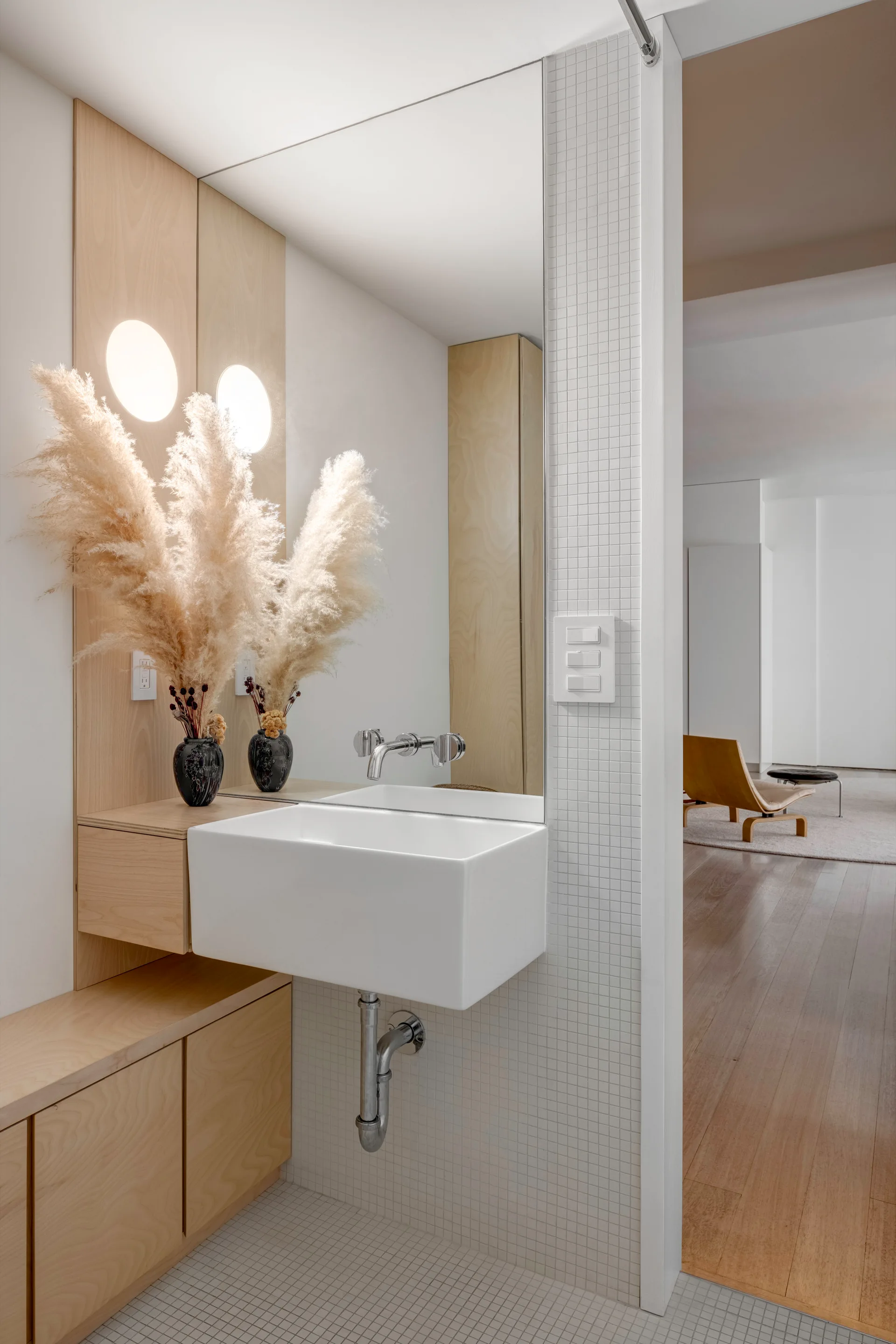 A minimalist bathroom features a white wall-mounted sink, light wood cabinetry, white square grid wall tiles, and a large mirror reflecting pampas grass.