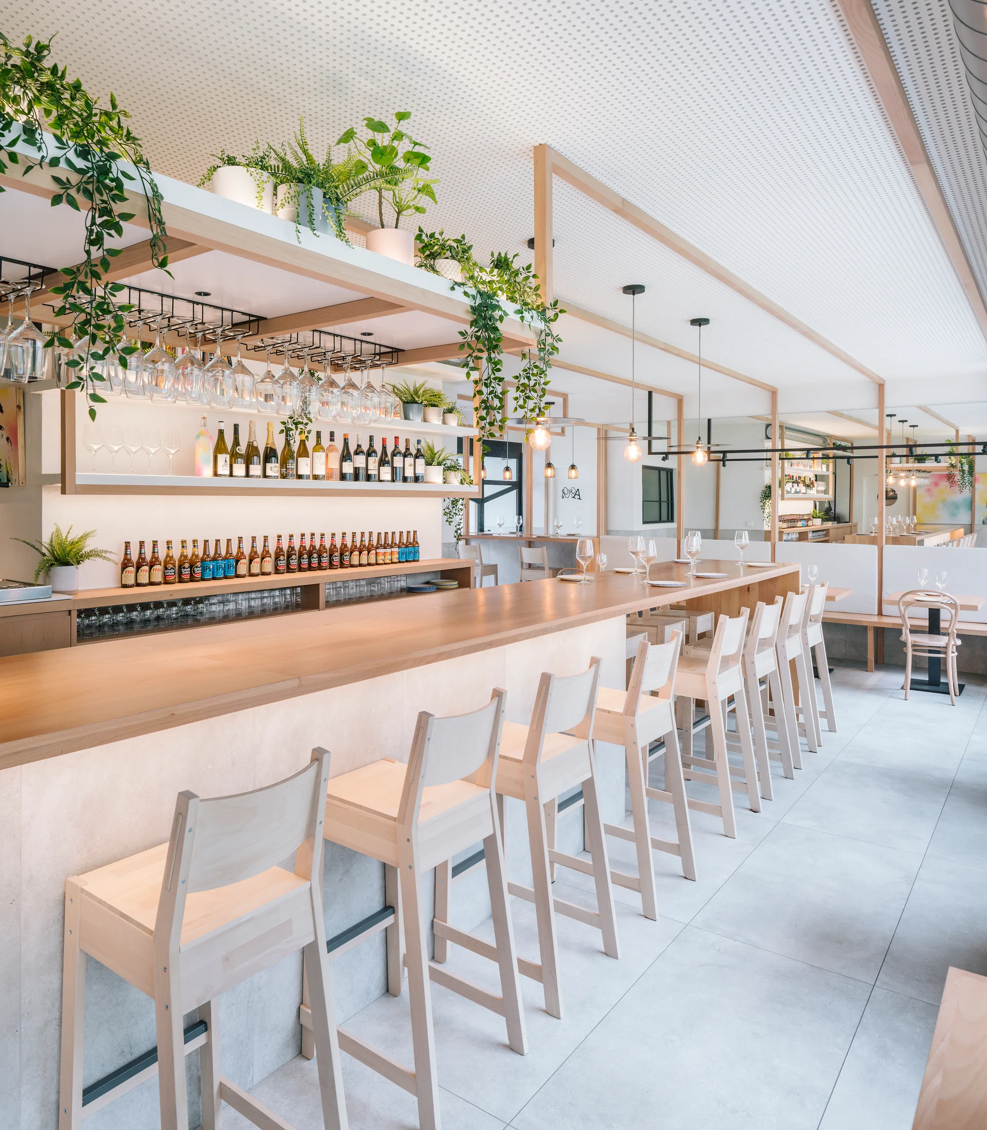 A minimalist restaurant bar features a light wood counter lined with matching wooden stools, open shelves filled with bottles, and hanging potted ivy.
