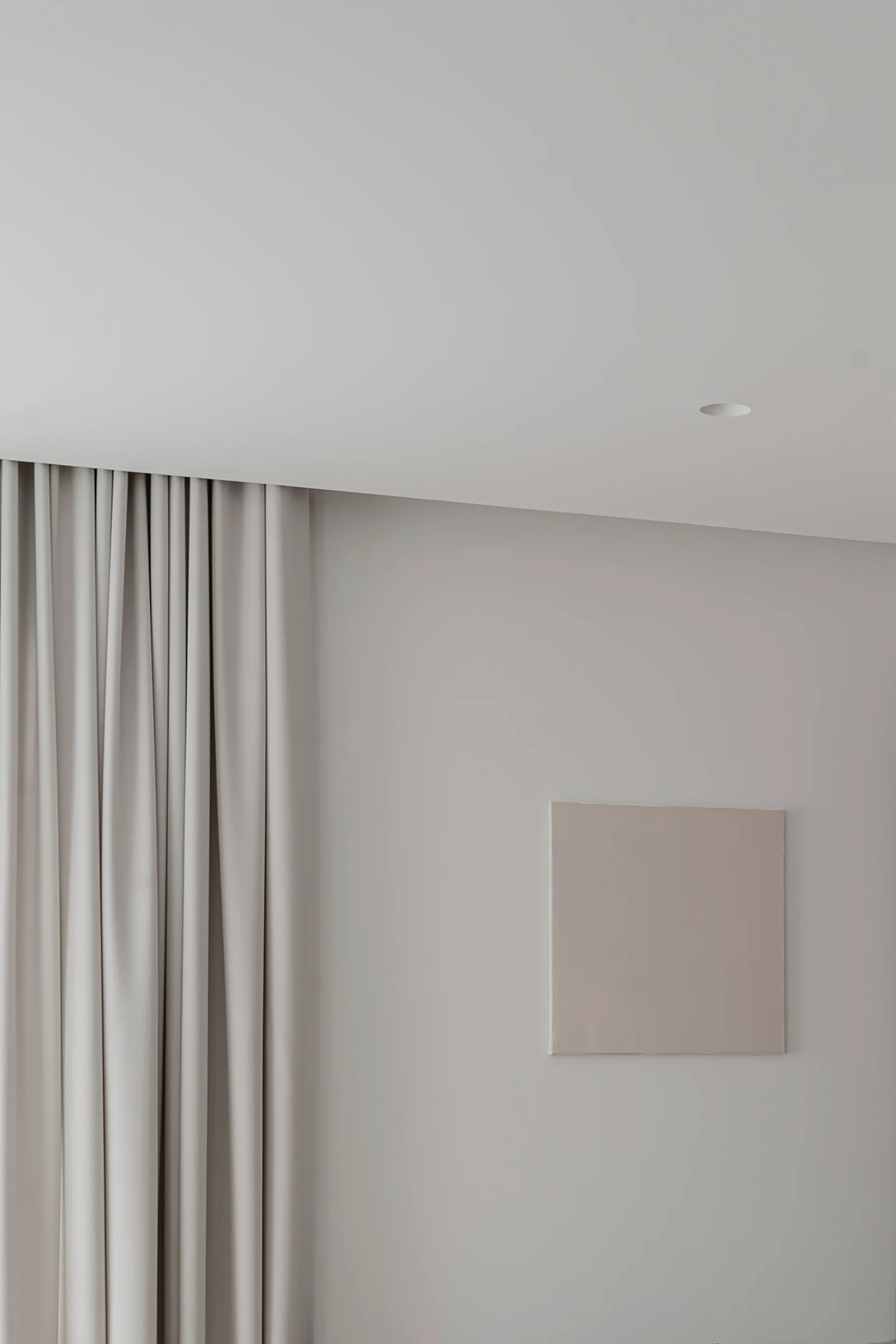 A minimalist beige wall featuring pleated floor-to-ceiling greige curtains, a recessed ceiling light, and a square canvas in a matching neutral tone.
