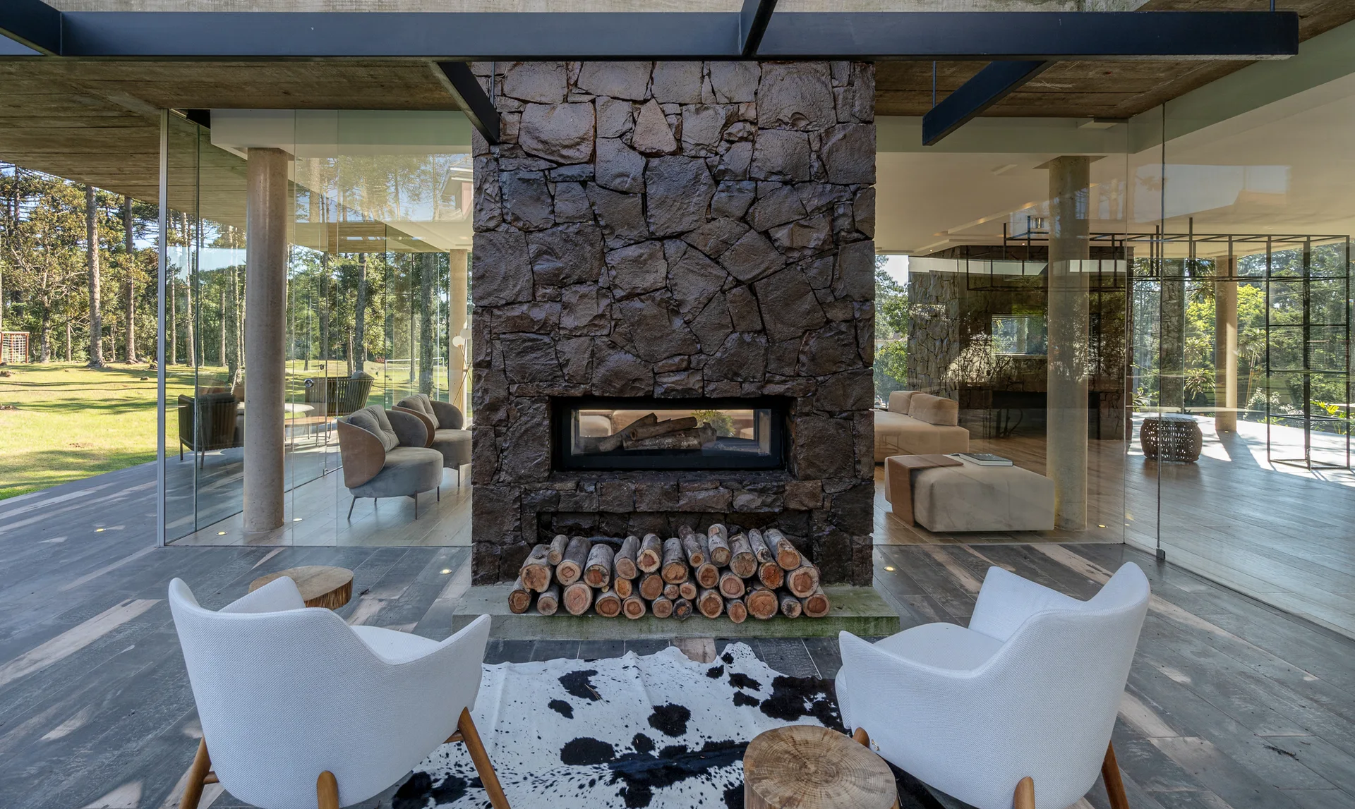A modern living room featuring floor-to-ceiling glass walls, a double-sided dark stone fireplace, white armchairs, and a cowhide rug.