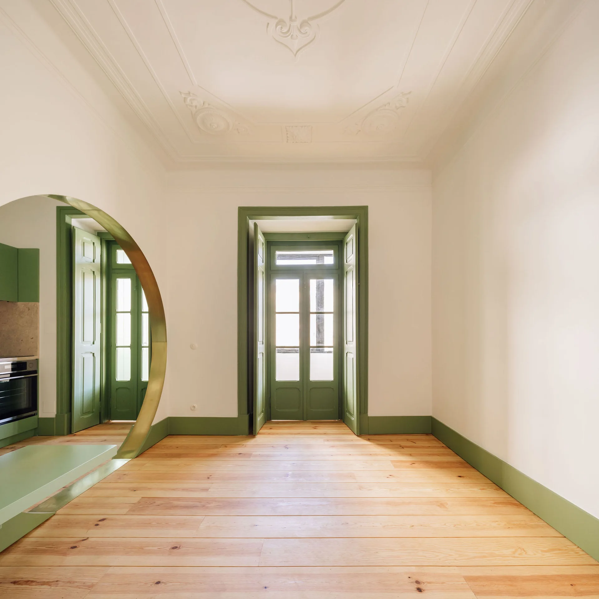 A bright room with light wood floors, white walls, sage green baseboards, and a circular gold archway leading into a kitchen.