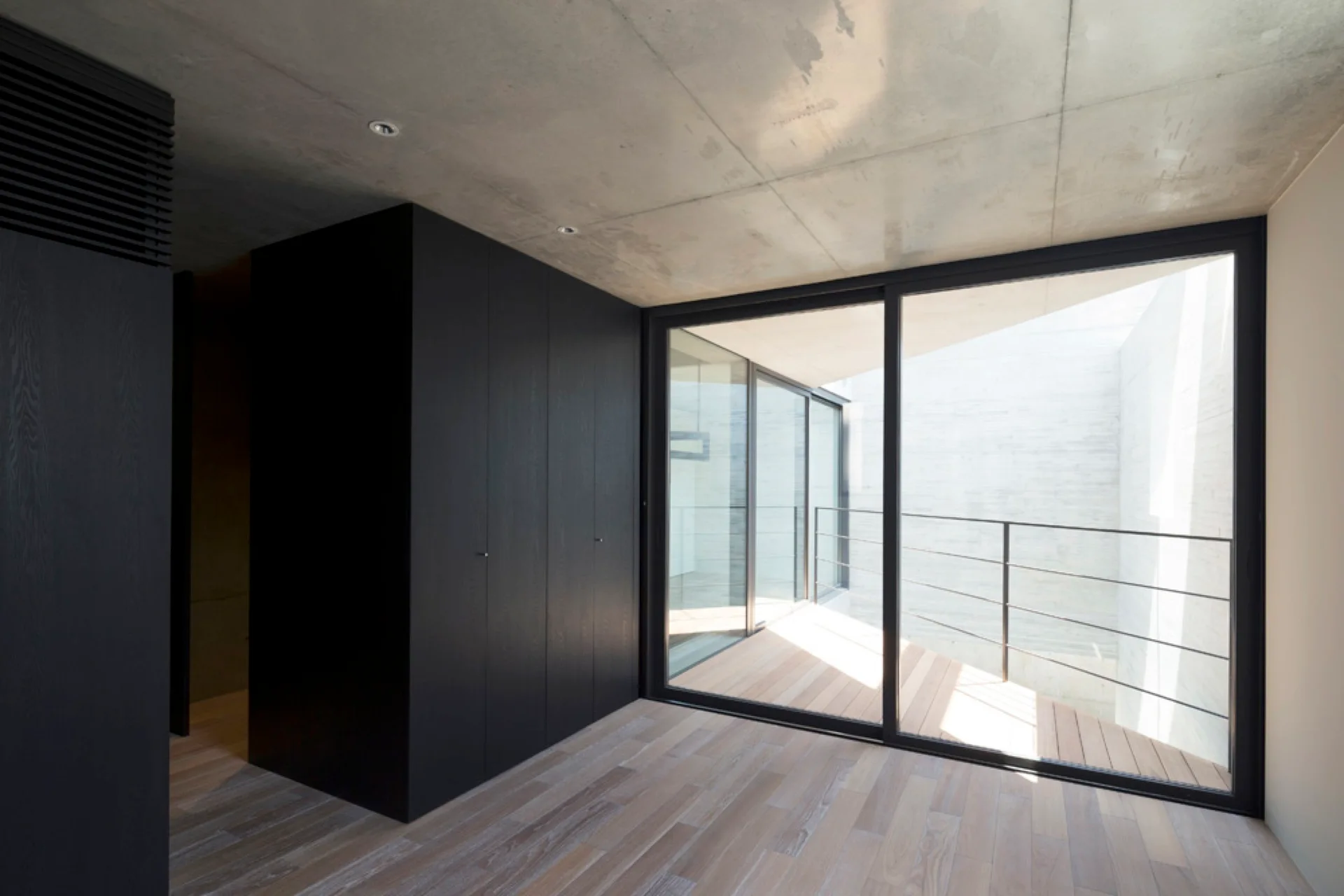 A minimalist room features light wood floors, an exposed concrete ceiling, a large black built-in cabinet, and a sliding glass door opening to a balcony.