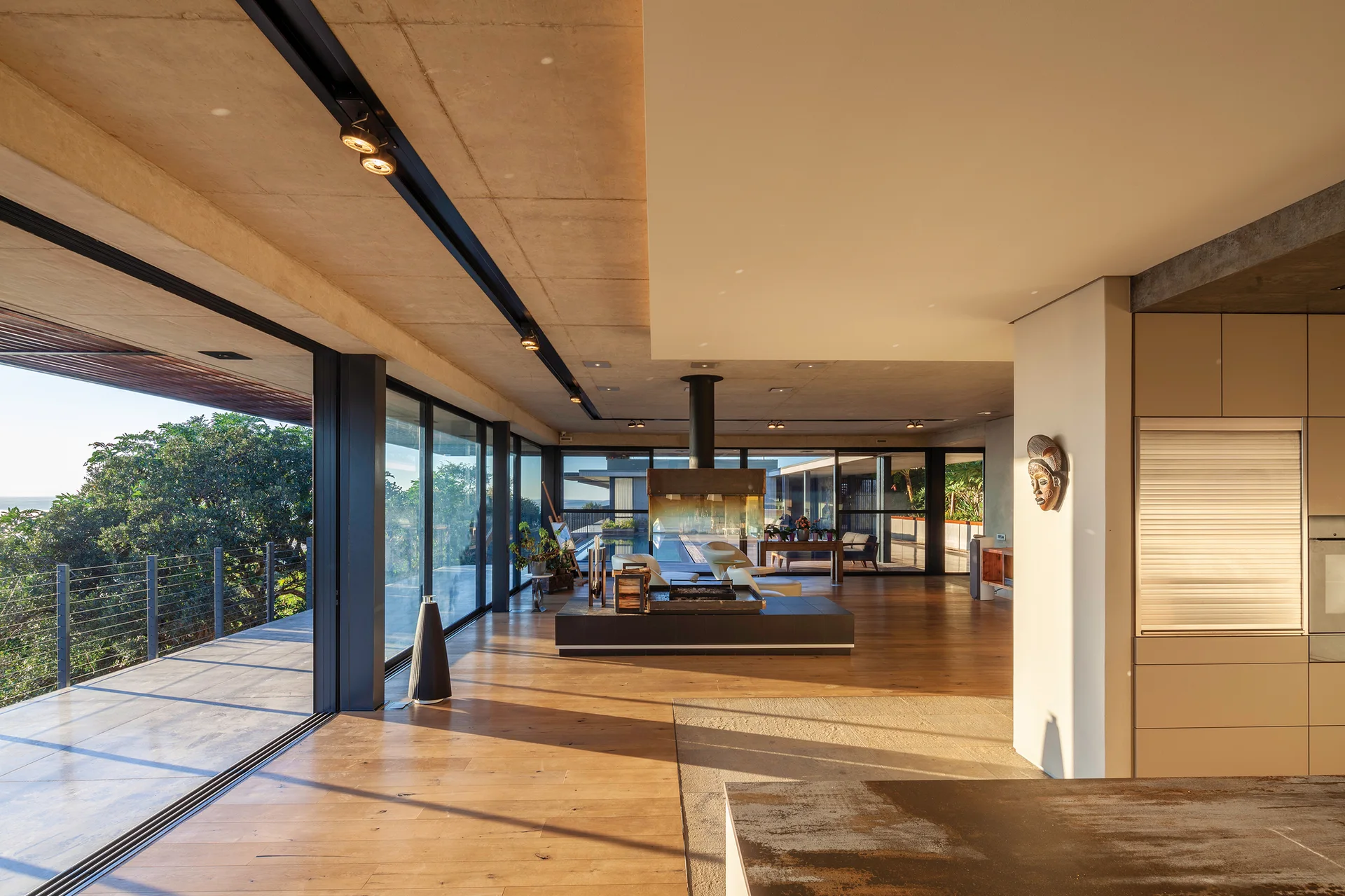An open-plan living room features an exposed concrete ceiling, wood floors, a central black fireplace, and floor-to-ceiling sliding glass doors.