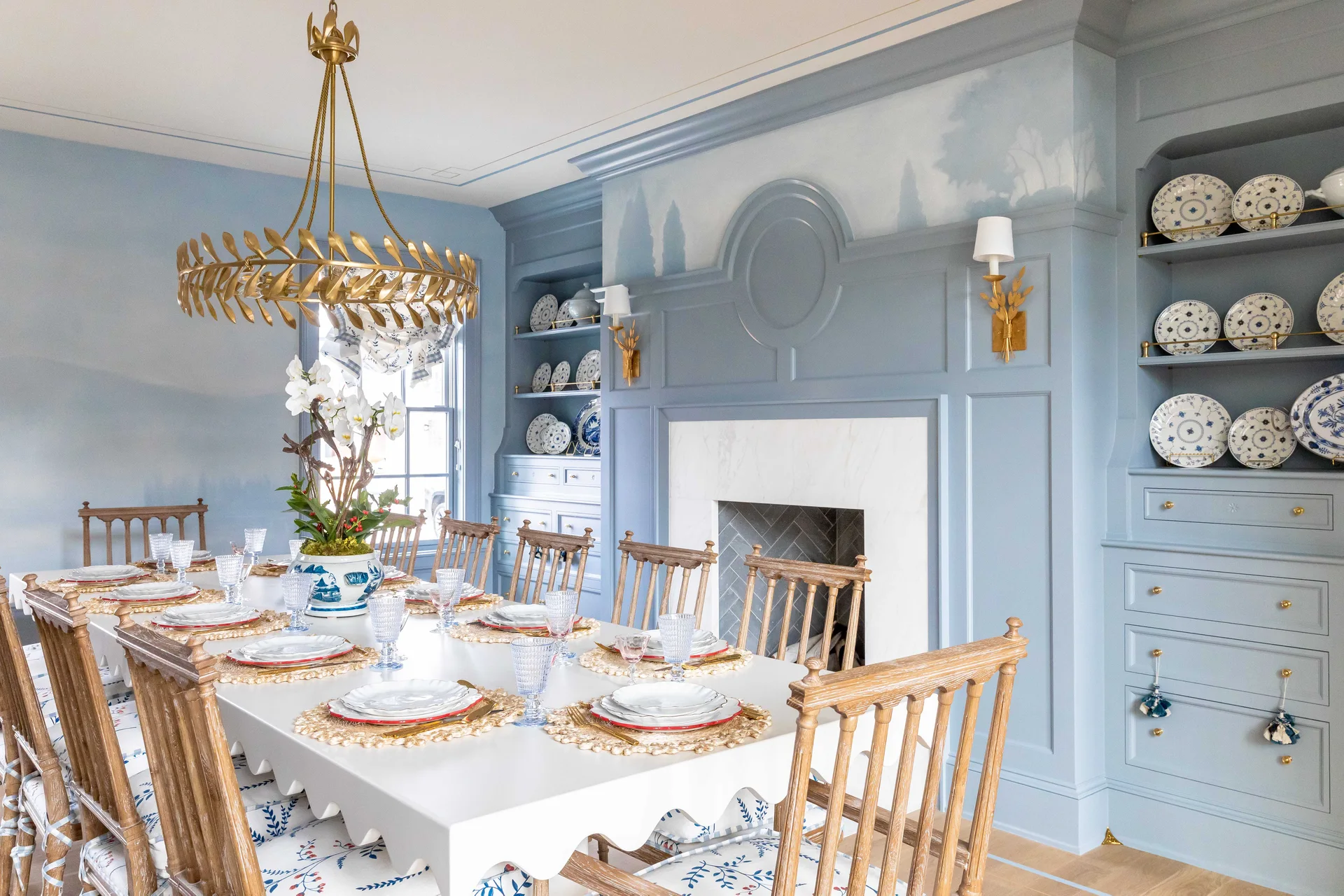A traditional dining room features a scalloped white table, wooden spindle chairs, a gold chandelier, and light blue built-in cabinetry surrounding a fireplace.
