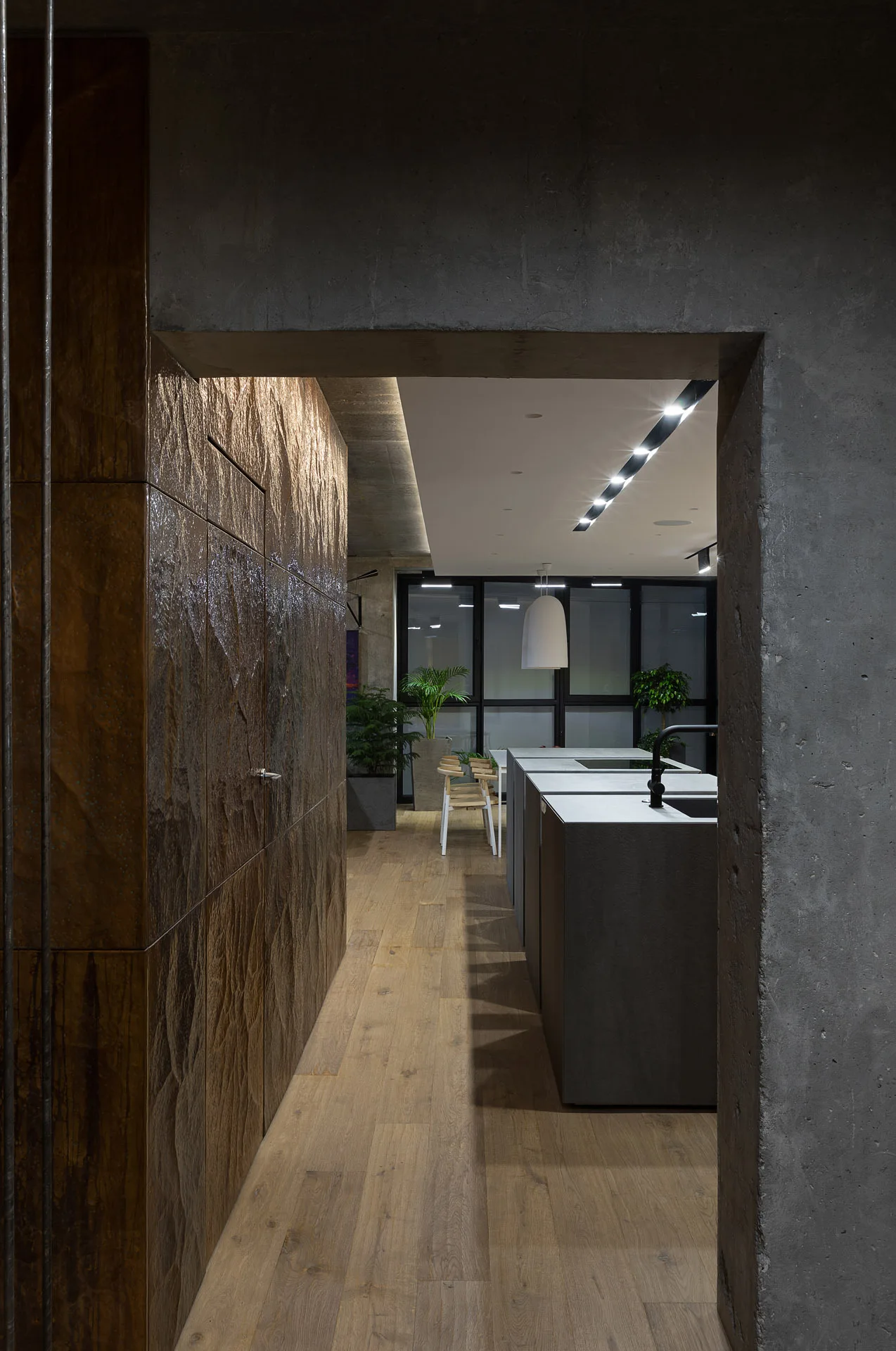 A view through a raw concrete doorway into a modern kitchen with a textured brown wall, a gray kitchen island, and light wood bar stools.