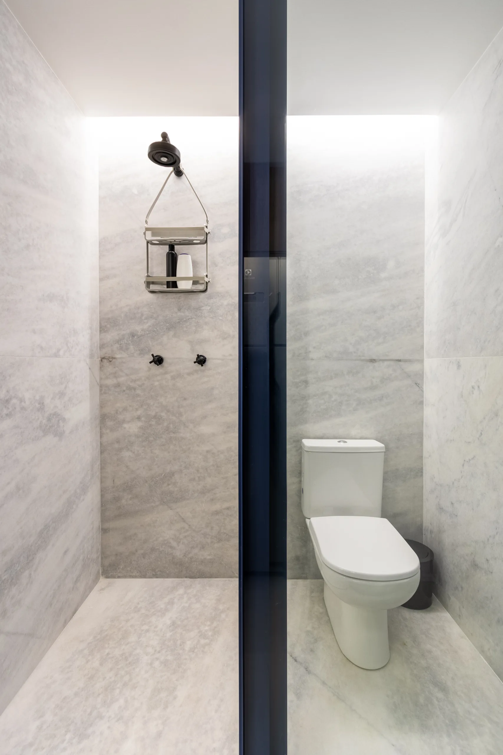 A modern bathroom features veined grey marble walls and floors, a white toilet, and a shower area separated by a deep blue glass partition.