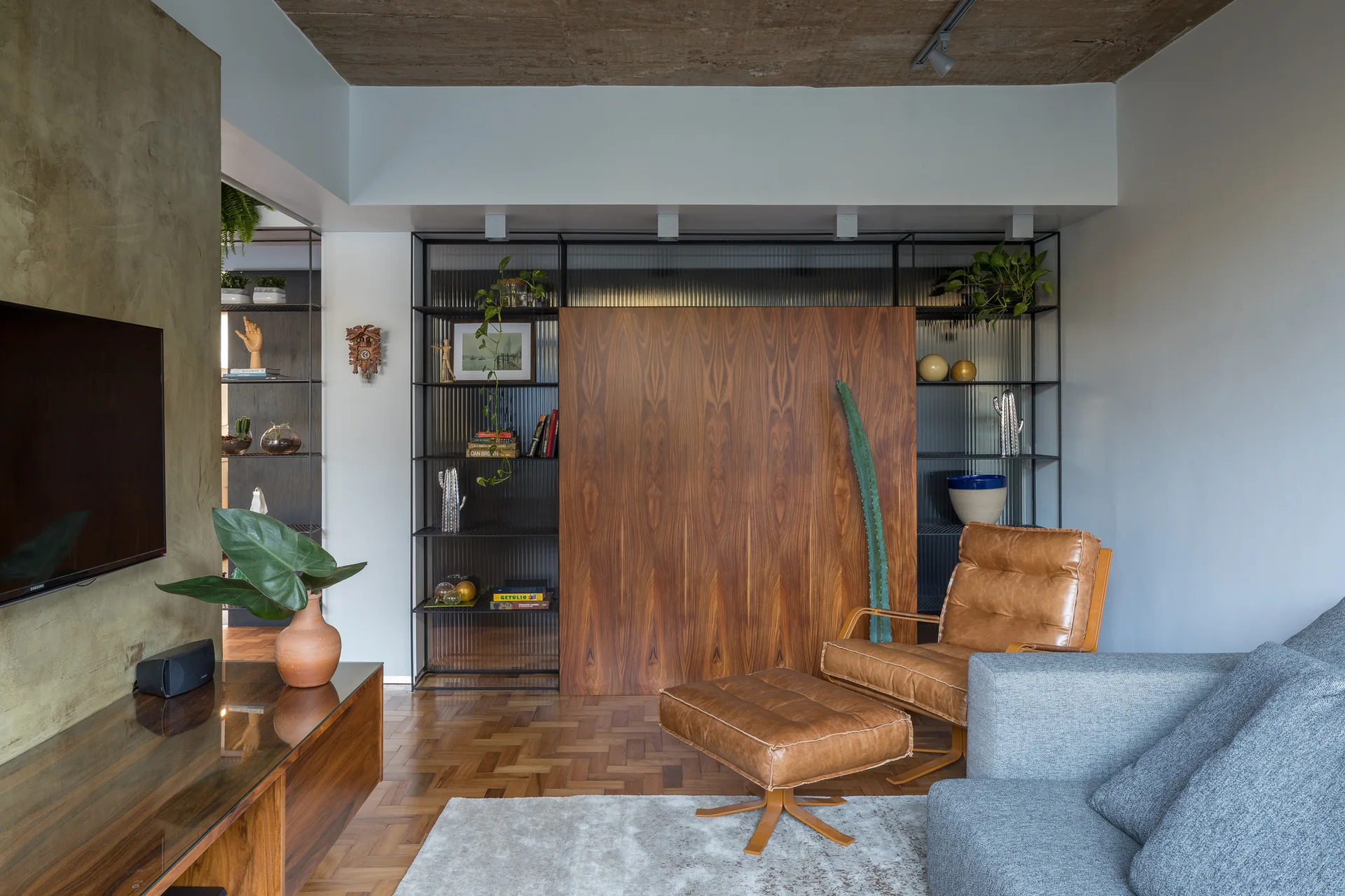 A contemporary living room features a tan leather armchair, parquet flooring, and a metal shelving unit backed by fluted glass with a central wood panel.