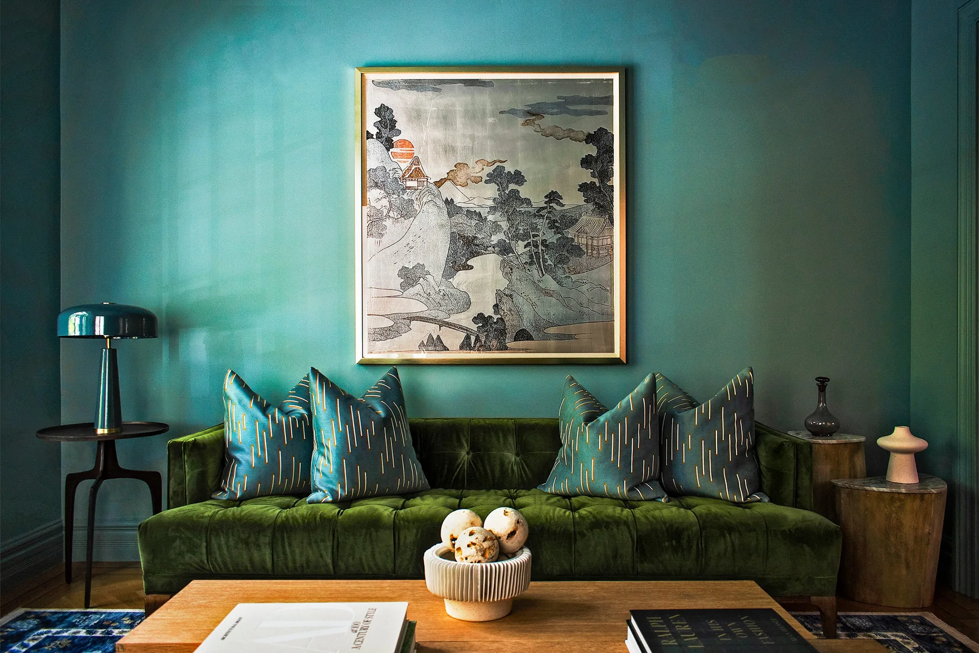 A dark green tufted velvet sofa sits against a teal wall beneath a gold-framed landscape painting in a living room.