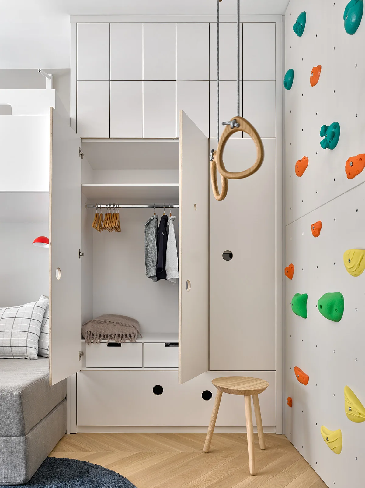 A kids room with a built-in white closet, hanging gymnastic rings, a wooden stool, and a climbing wall with colorful holds on herringbone floors.