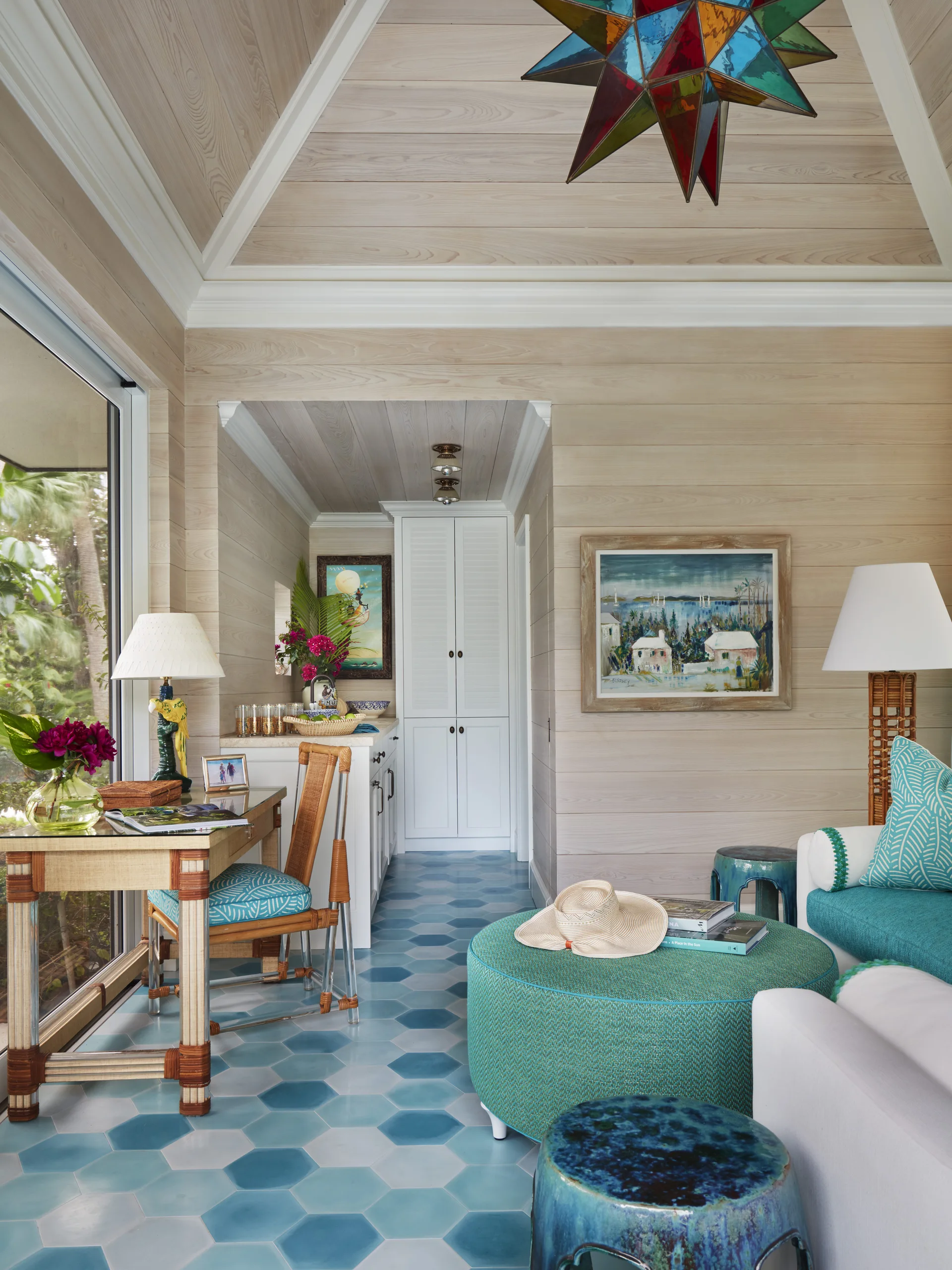 A sunlit coastal living room features light wood walls, blue hexagonal floor tiles, a writing desk, and a large teal ottoman.