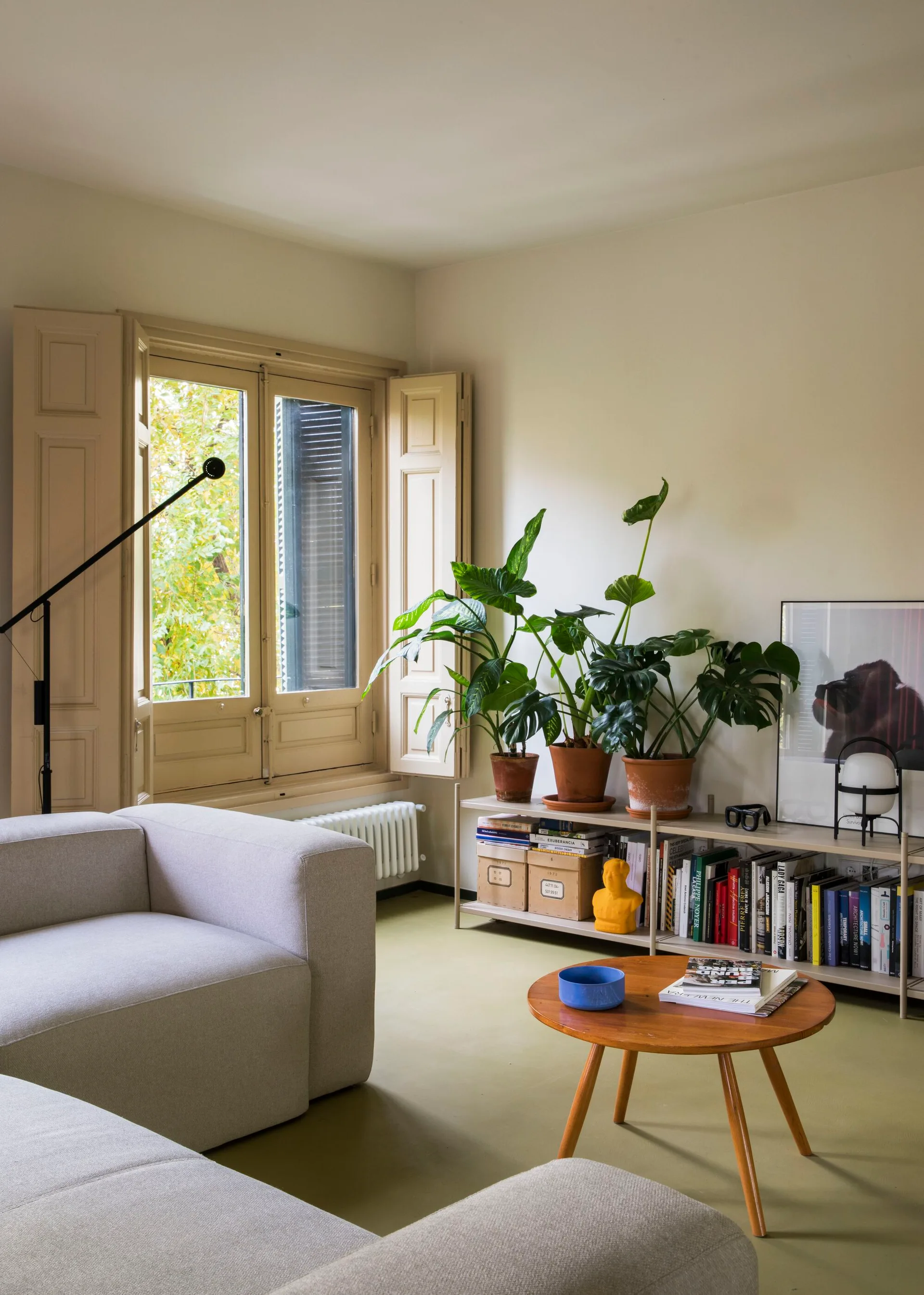A living room with a beige fabric sofa, a round wood coffee table, green flooring, and a low shelf filled with books and potted plants.