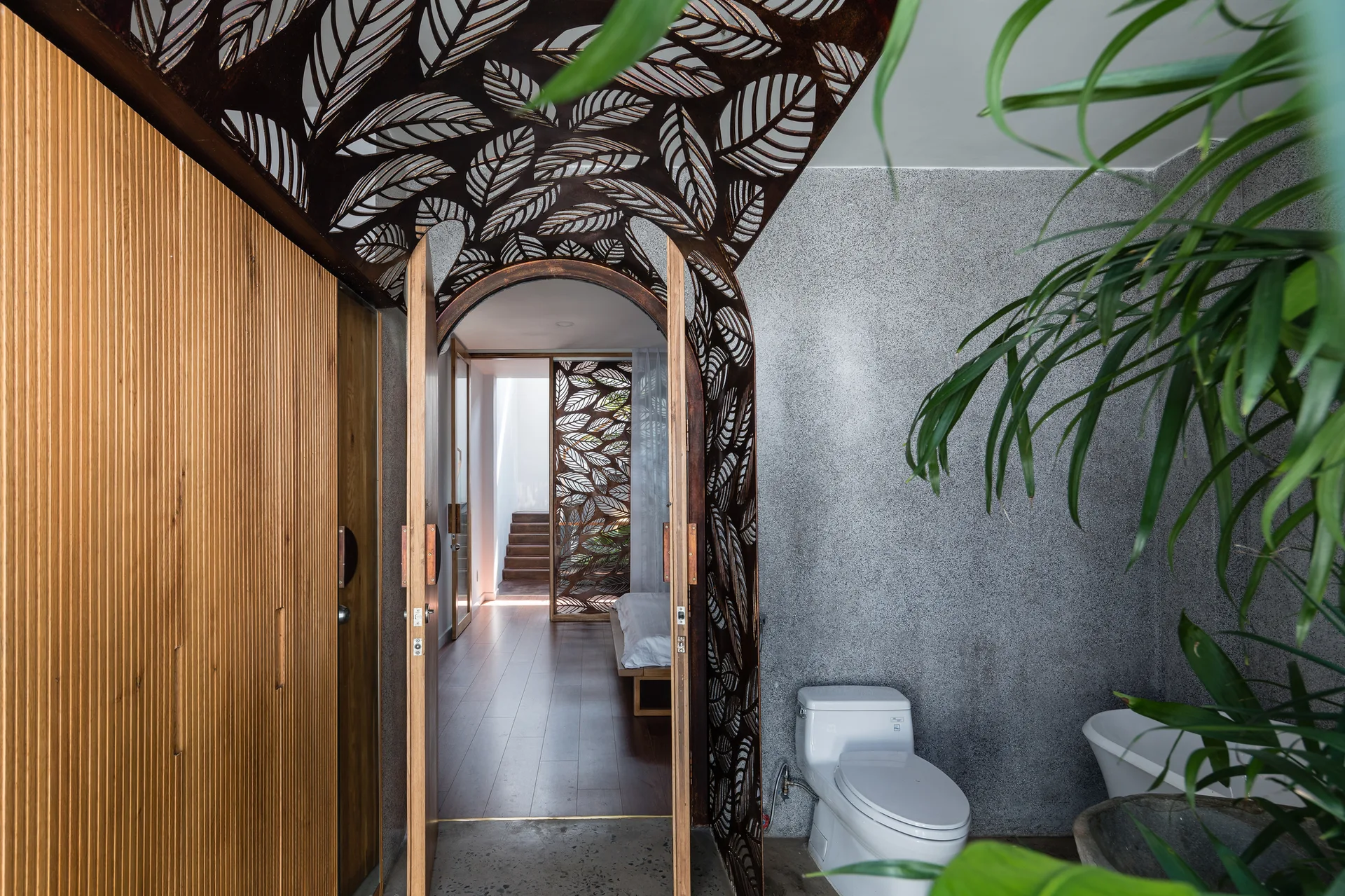 A modern bathroom features a decorative laser-cut leaf pattern metal archway, textured grey stone walls, and a vertical wood slat partition.