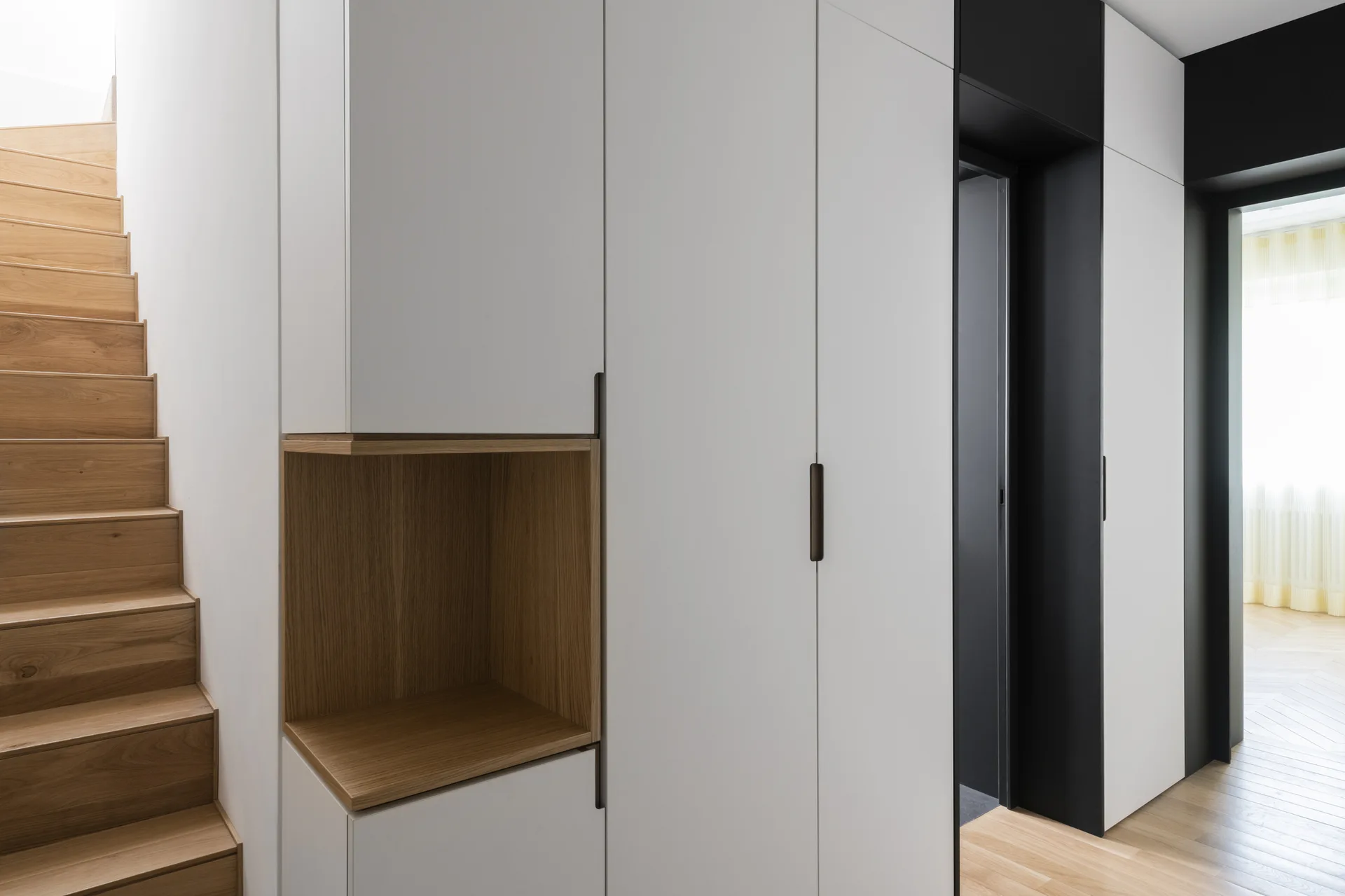 Modern white built-in cabinetry with a wood-toned alcove stands next to a wooden staircase and a dark passage.