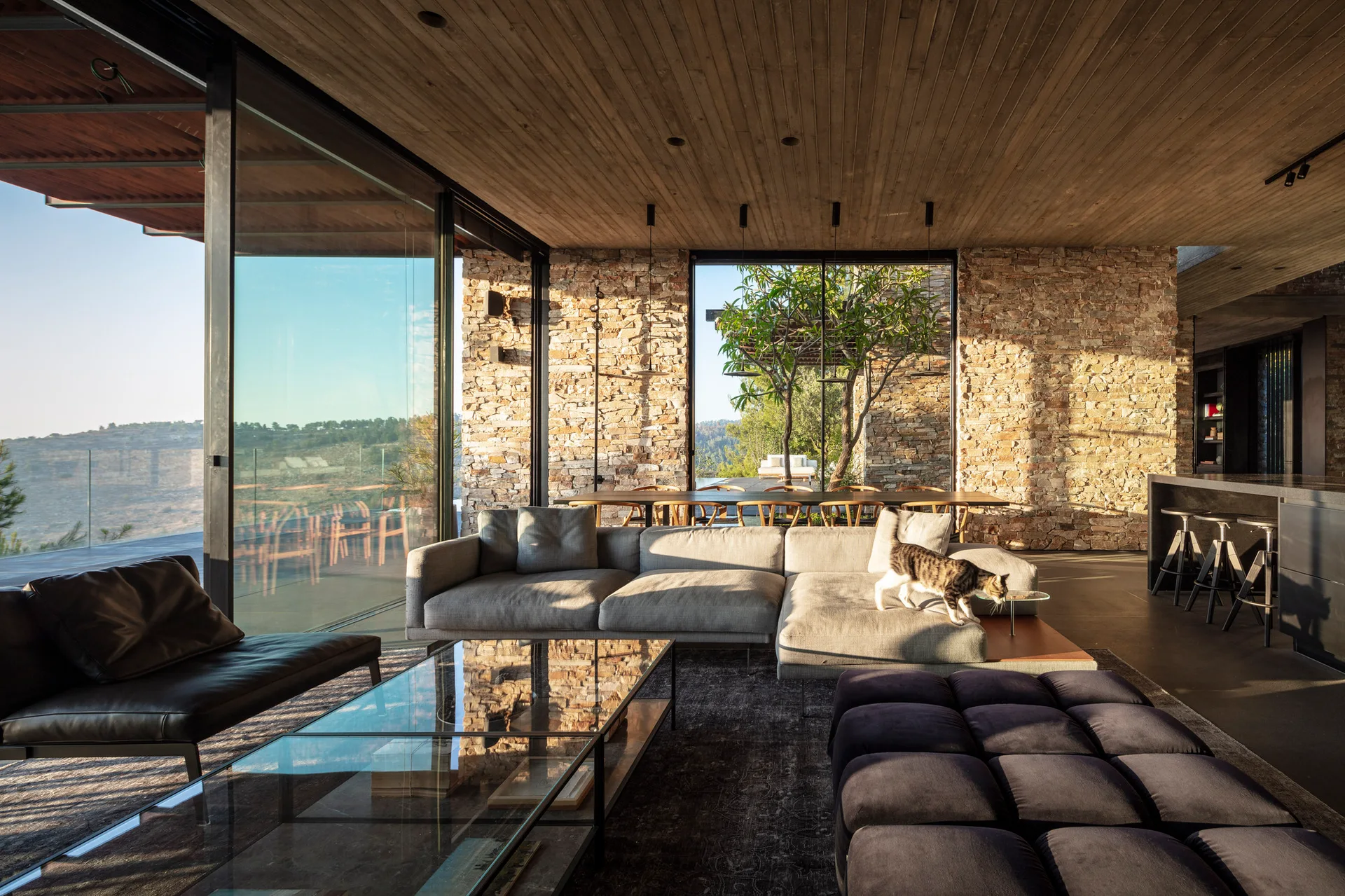 A modern open-plan living room features a grey modular sofa, a glass coffee table, exposed stone walls, and a wood plank ceiling.