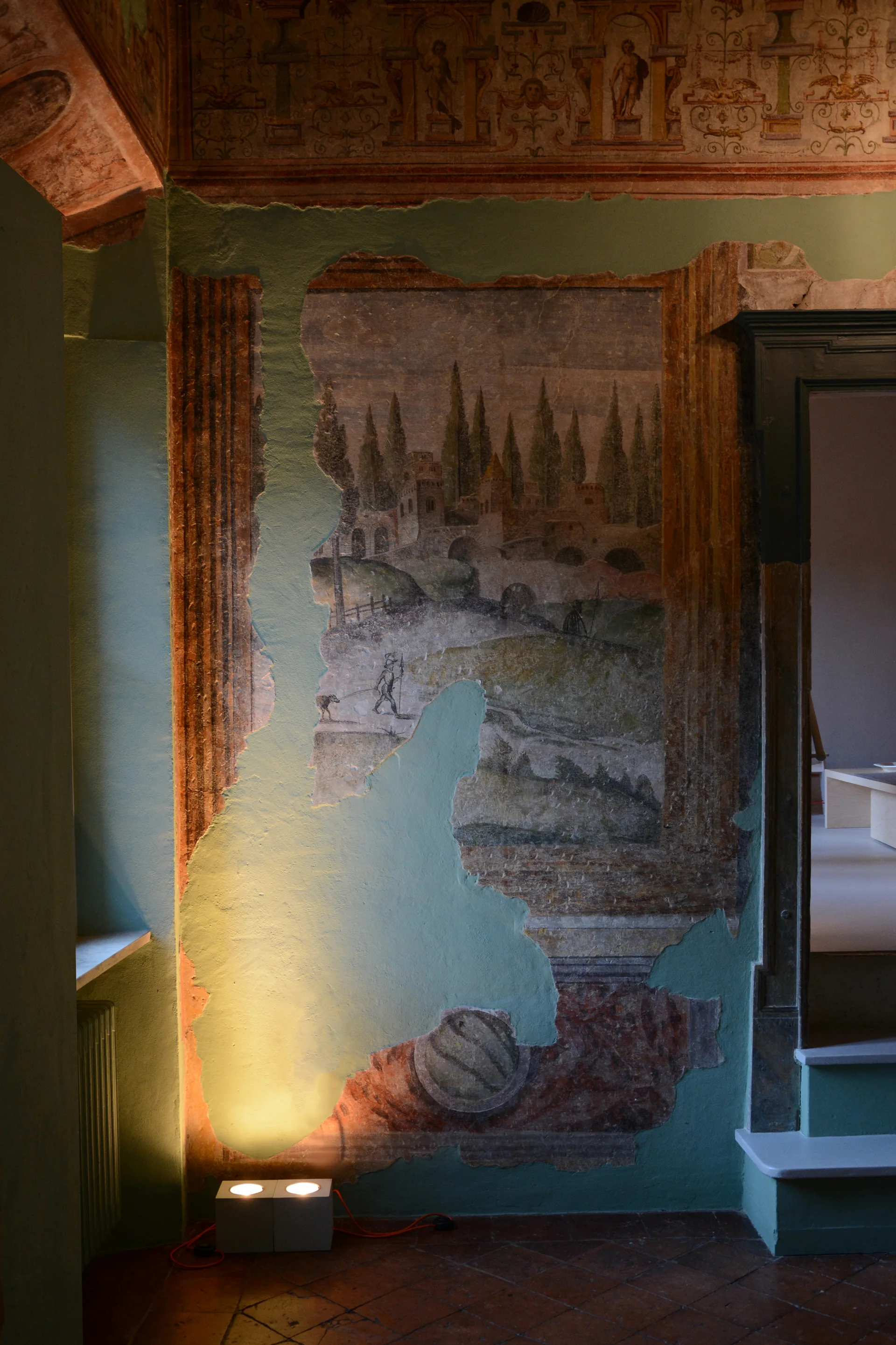 An antique landscape fresco is revealed through a distressed sage green plaster wall, lit by a dual box floor lamp next to a doorway.