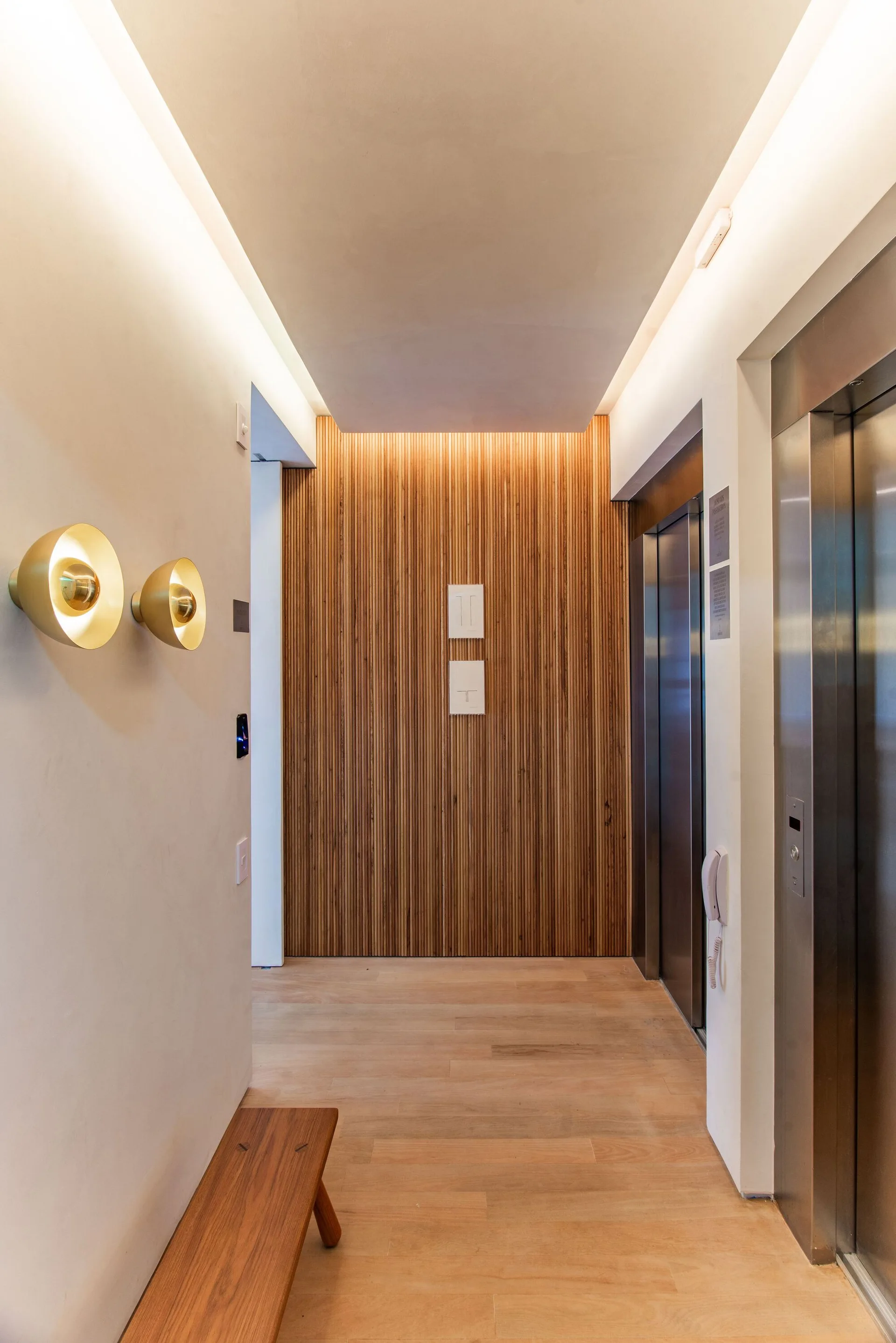 A modern elevator lobby featuring a slatted wood accent wall, recessed cove lighting, and dual round brass sconces on an off-white wall.