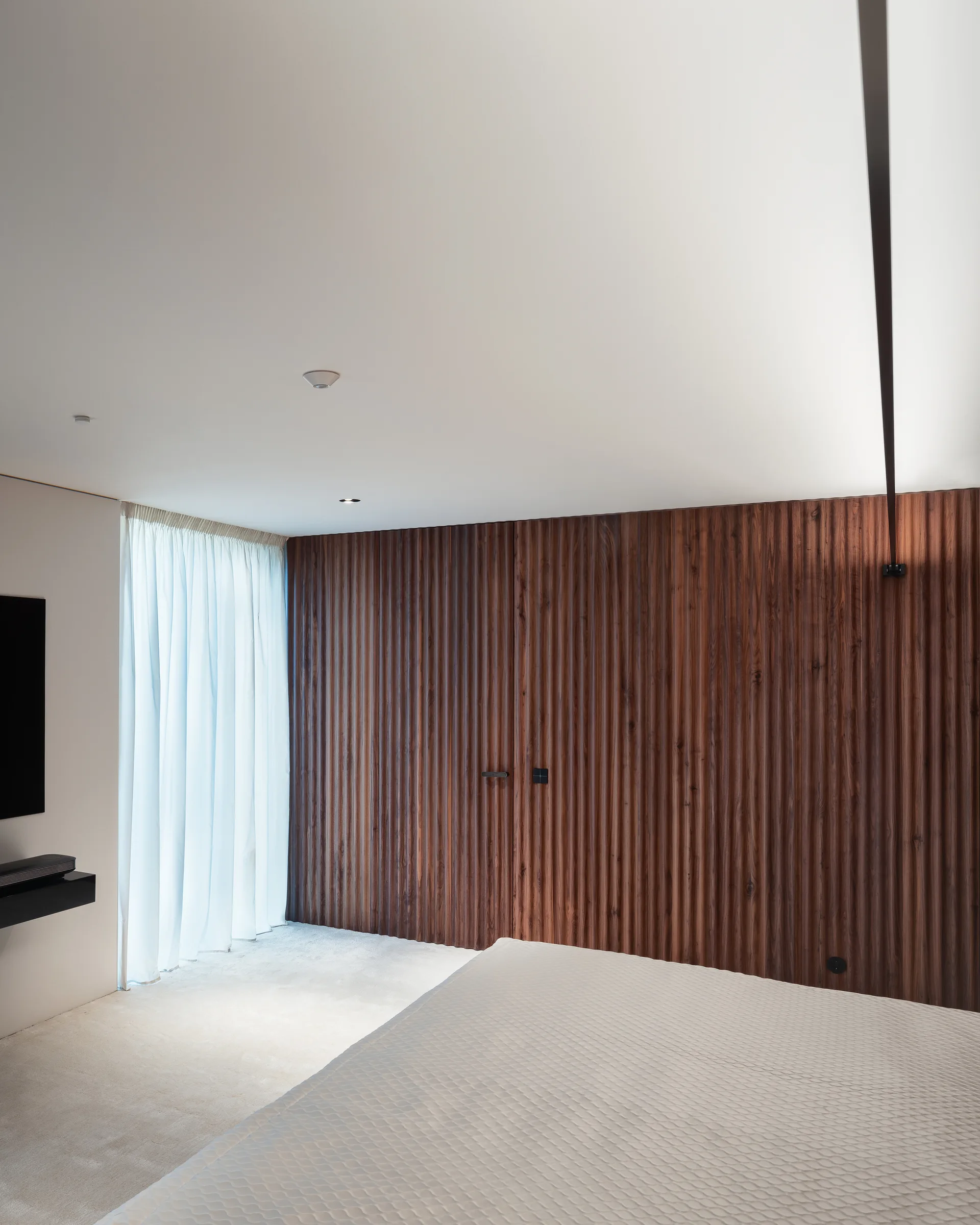 A minimalist bedroom featuring a textured dark wood fluted wall panel, a quilted white bed coverlet, and sheer white curtains.