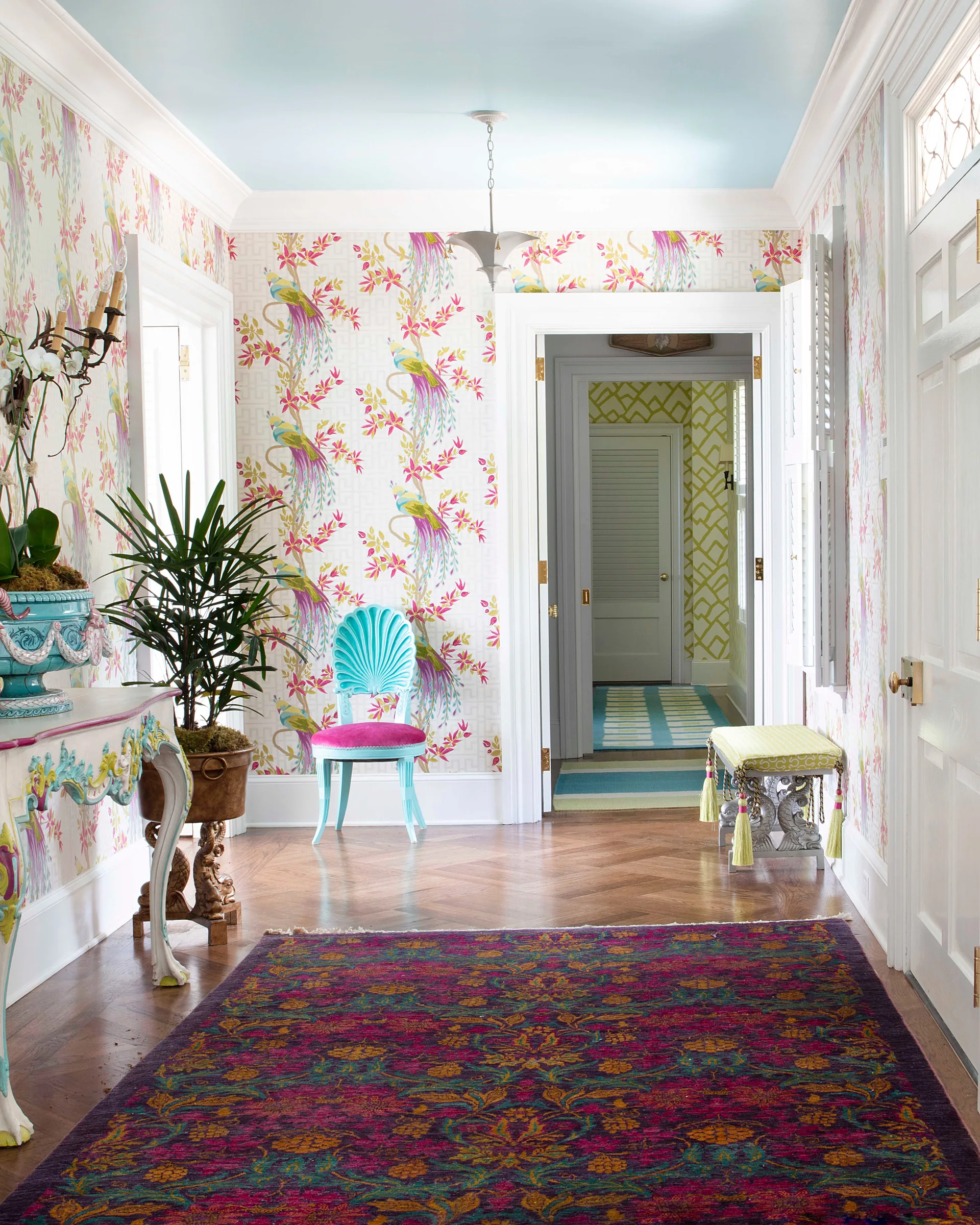 A vibrant entryway features floral bird wallpaper, a light blue ceiling, a turquoise shell chair, and herringbone wood flooring.