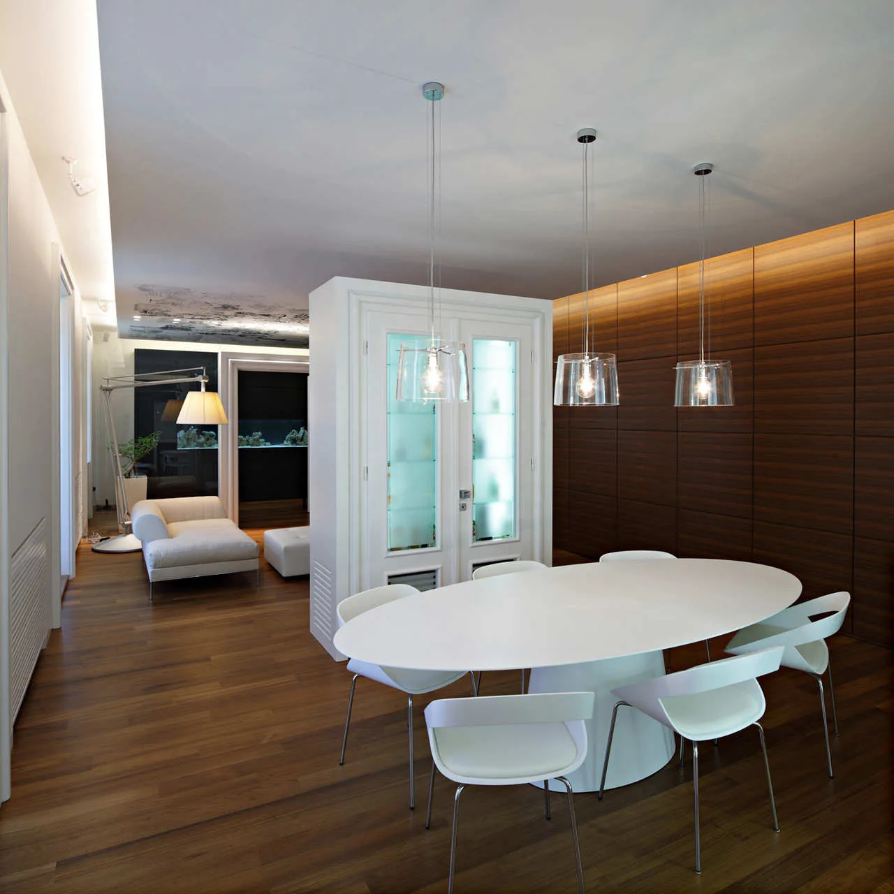 A modern dining room with a white oval table and chairs, clear glass pendant lights, a white display cabinet, and a dark wood paneled wall.