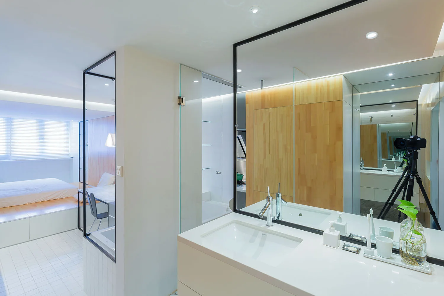 A modern white bathroom vanity with a black-framed mirror next to a glass partition, showing a bedroom with wood paneling in the background.