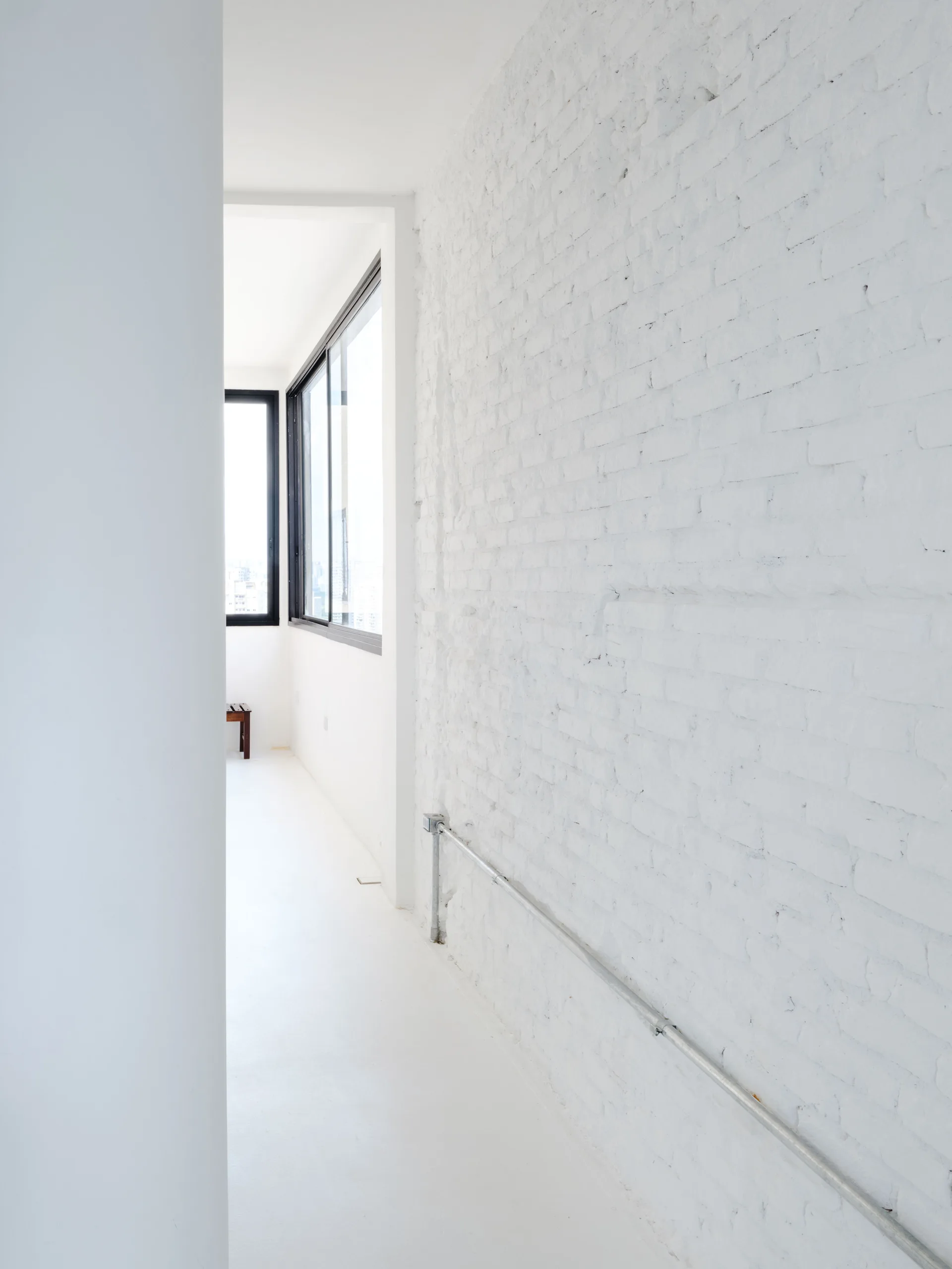 A bright white minimalist hallway features an exposed painted brick wall, a metal conduit pipe, and black-framed windows at the far end.