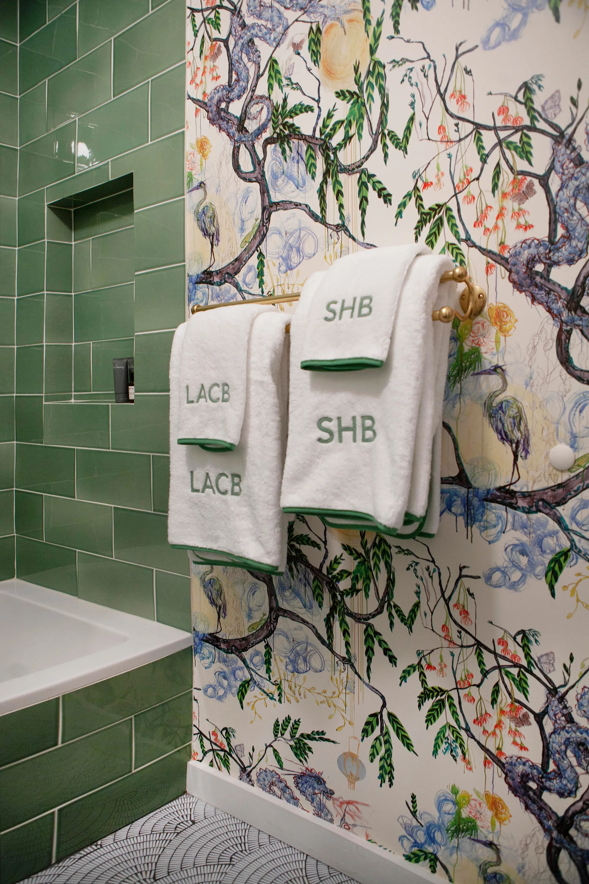 A bathroom wall with green subway tiles and a recessed niche next to botanical wallpaper and white monogrammed towels on a brass rack.