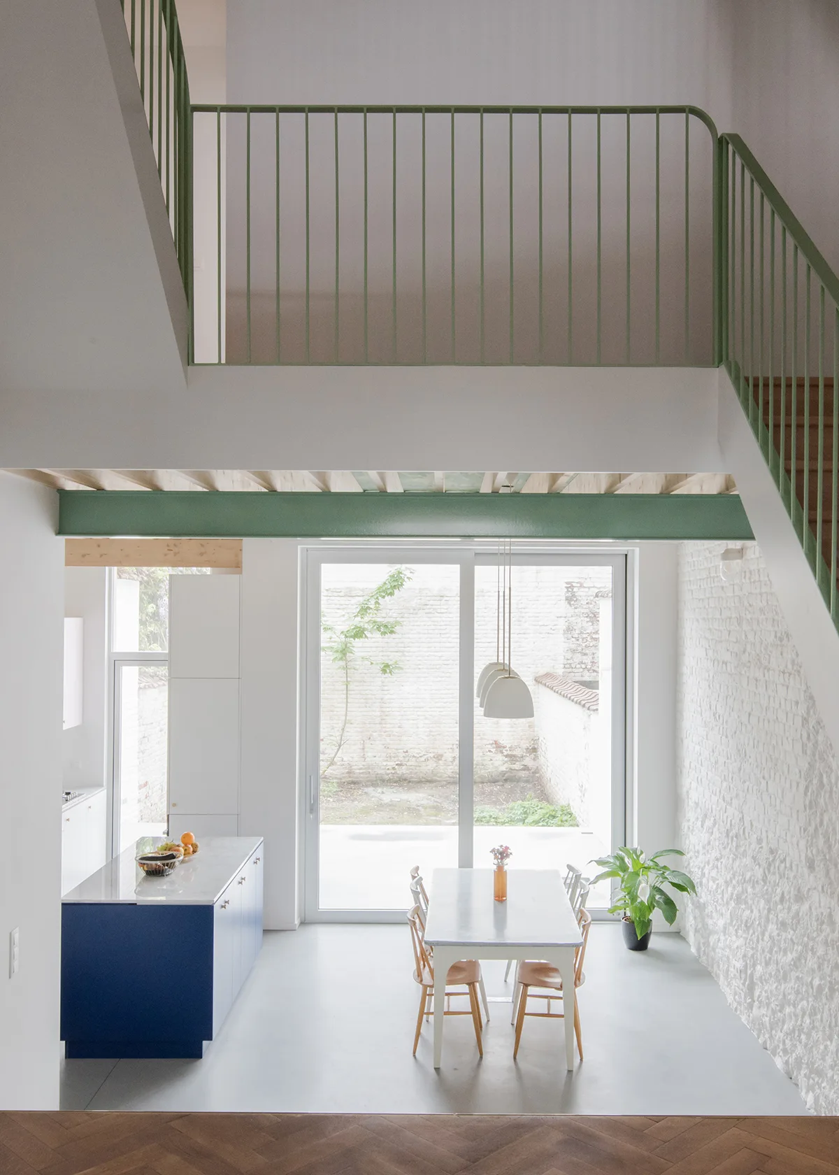 A high-angle view of an open-plan dining space featuring a green metal staircase railing, a blue kitchen island, and a textured white brick wall.