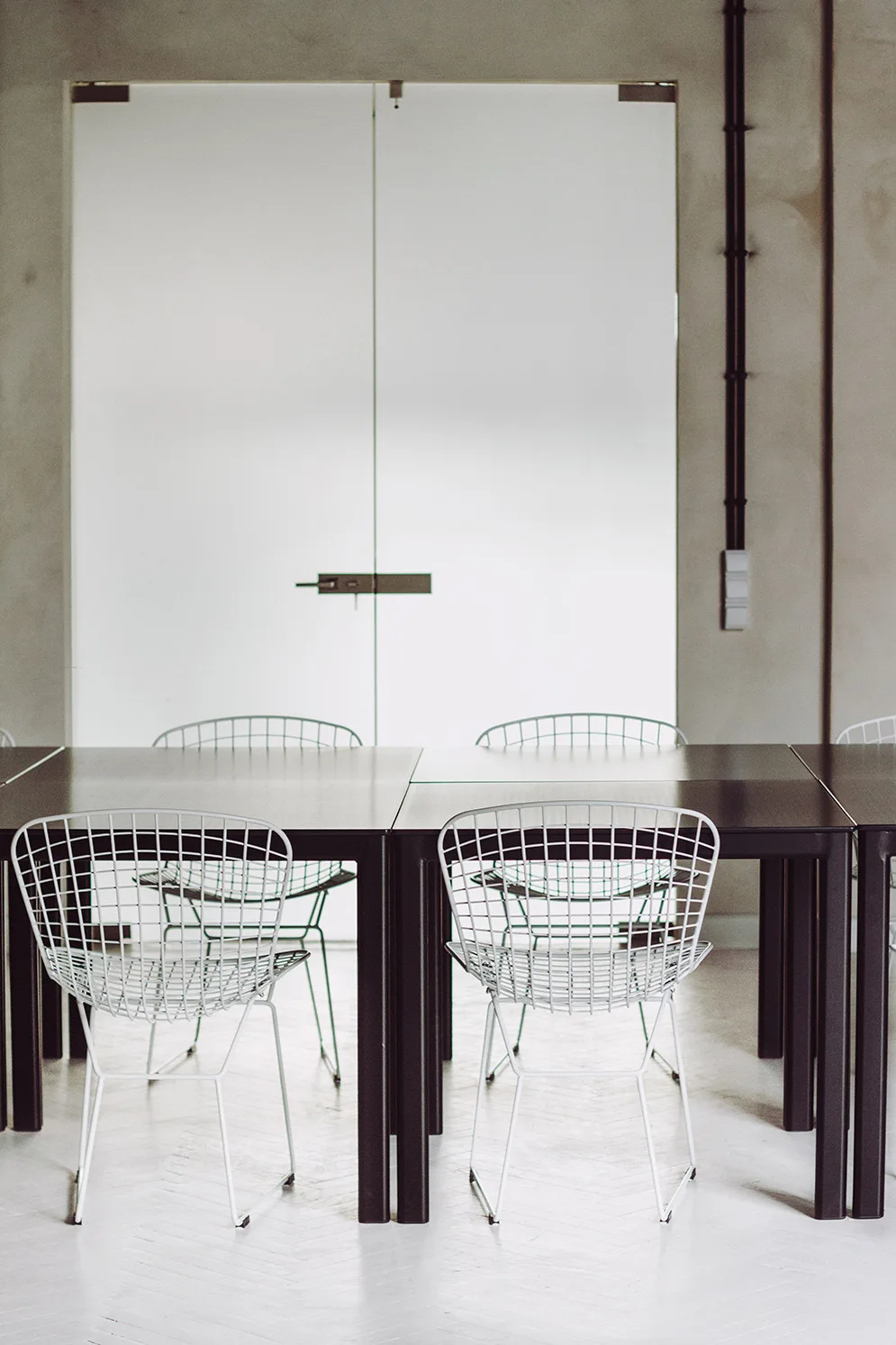 A minimalist dining area features dark wooden tables paired with white wire-grid chairs on a light herringbone floor.