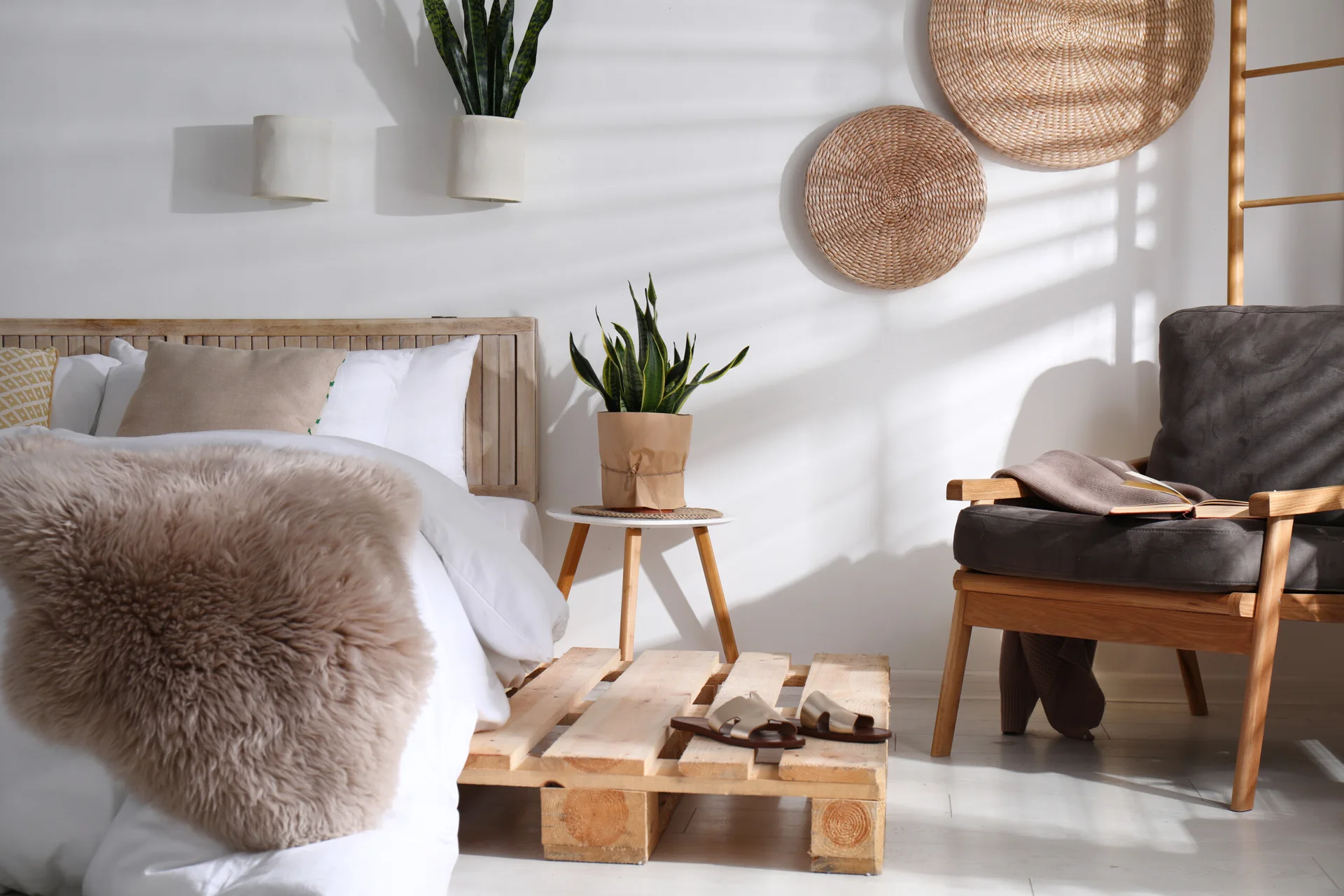 A minimalist bedroom featuring a bed with a wooden headboard, a dark armchair, circular woven wall decor, and potted snake plants.