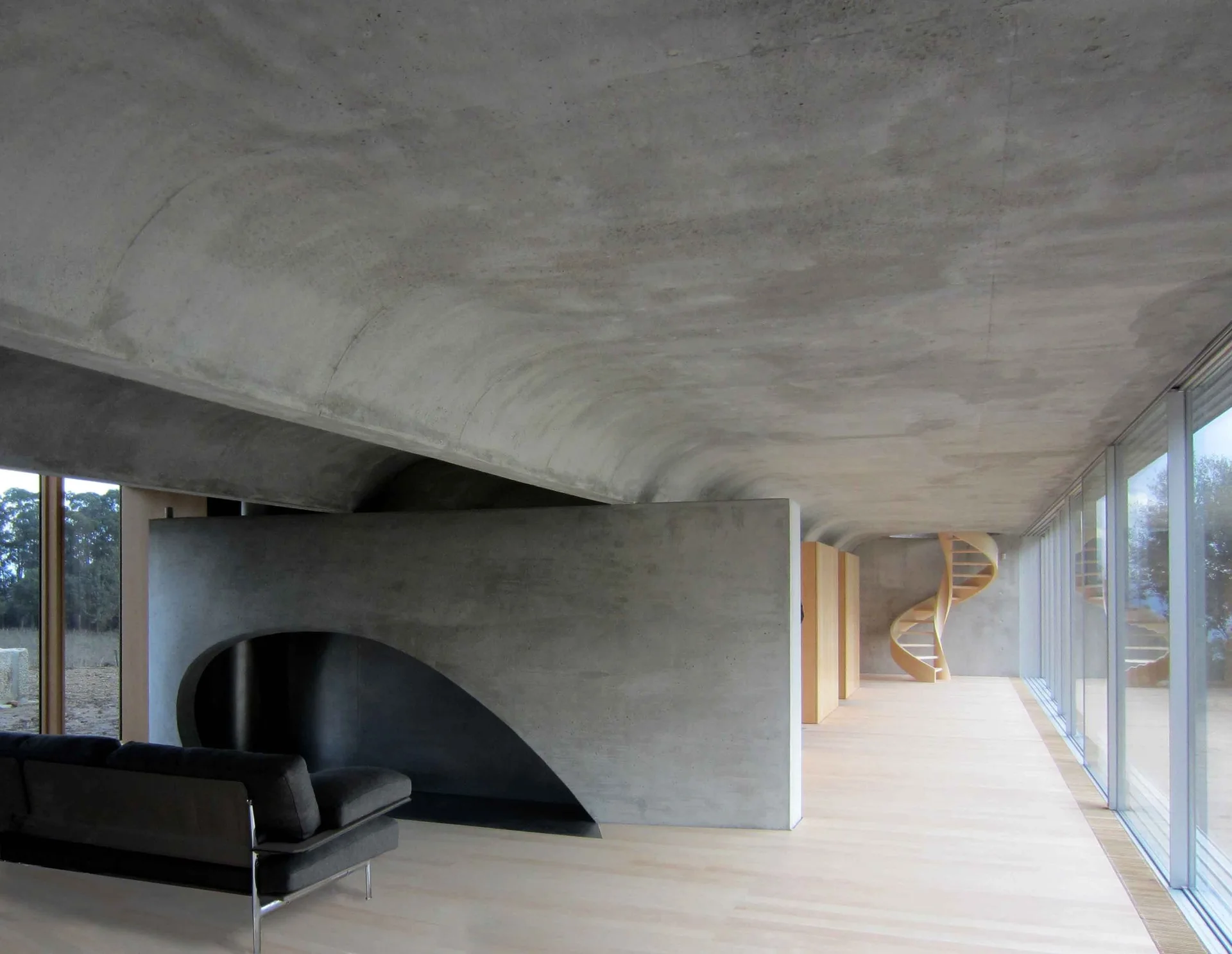 A modern minimalist room featuring a curved concrete ceiling, a concrete partition wall with a cutout, wood flooring, and a spiral staircase.