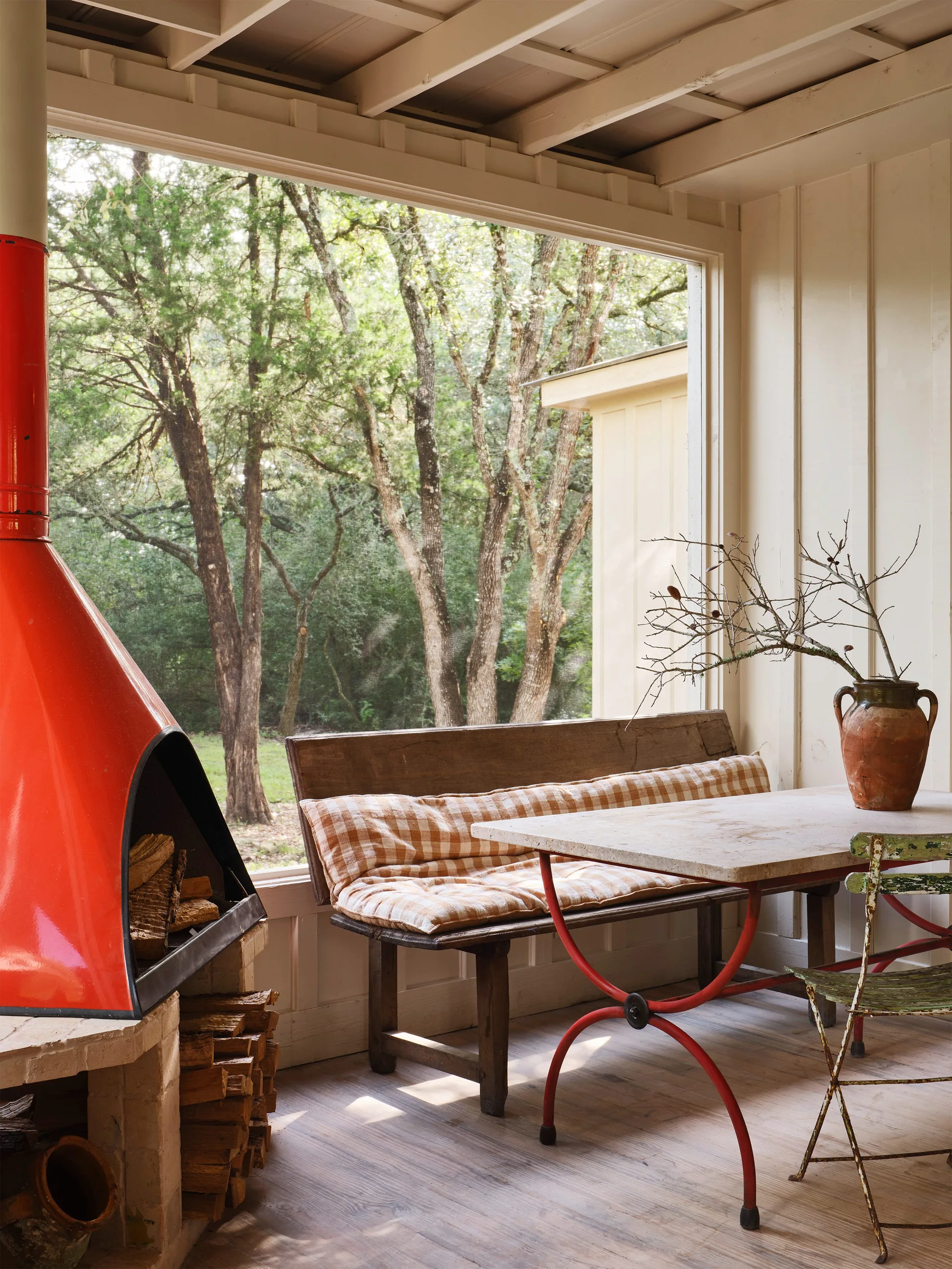 A rustic covered porch featuring a bright red metal fireplace, a wooden bench with a checkered cushion, and a dining table next to a large window.
